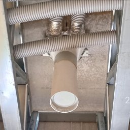 JP TANI DOM SPÓŁKA Z OGRANICZONĄ ODPOWIEDZIALNOŚCIĄ - Widok z dołu na instalację wentylacyjną w trakcie montażu, z widocznymi rurami karbowanymi, elementami izolacji z folii aluminiowej i konstrukcją szkieletową sufitu podwieszanego.