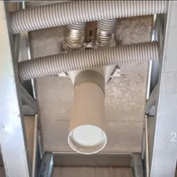 Widok z dołu na instalację wentylacyjną w trakcie montażu, z widocznymi rurami karbowanymi, elementami izolacji z folii aluminiowej i konstrukcją szkieletową sufitu podwieszanego.