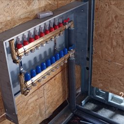 JP TANI DOM SPÓŁKA Z OGRANICZONĄ ODPOWIEDZIALNOŚCIĄ - Rozdzielacz ogrzewania podłogowego z mosiężnymi rurami i czerwono-niebieskimi zaworami, zamontowany na metalowej konstrukcji, widoczne fragmenty ściany z płyty OSB.