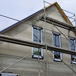 JP TANI DOM SPÓŁKA Z OGRANICZONĄ ODPOWIEDZIALNOŚCIĄ - Surowa elewacja domu w trakcie budowy, widoczne rusztowania, okna z roletami zewnętrznymi, szara struktura elewacji z widocznymi mocowaniami.