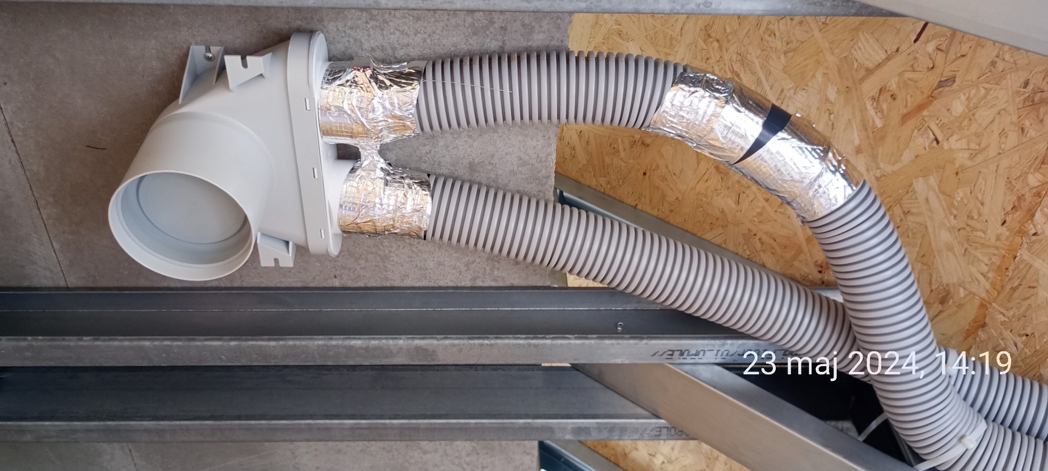 Dwie elastyczne rury wentylacyjne, jedna owinięta folią aluminiową i taśmą, podłączone do białej plastikowej złączki, zamocowane do sufitu z płyty OSB i metalowej konstrukcji szkieletowej.