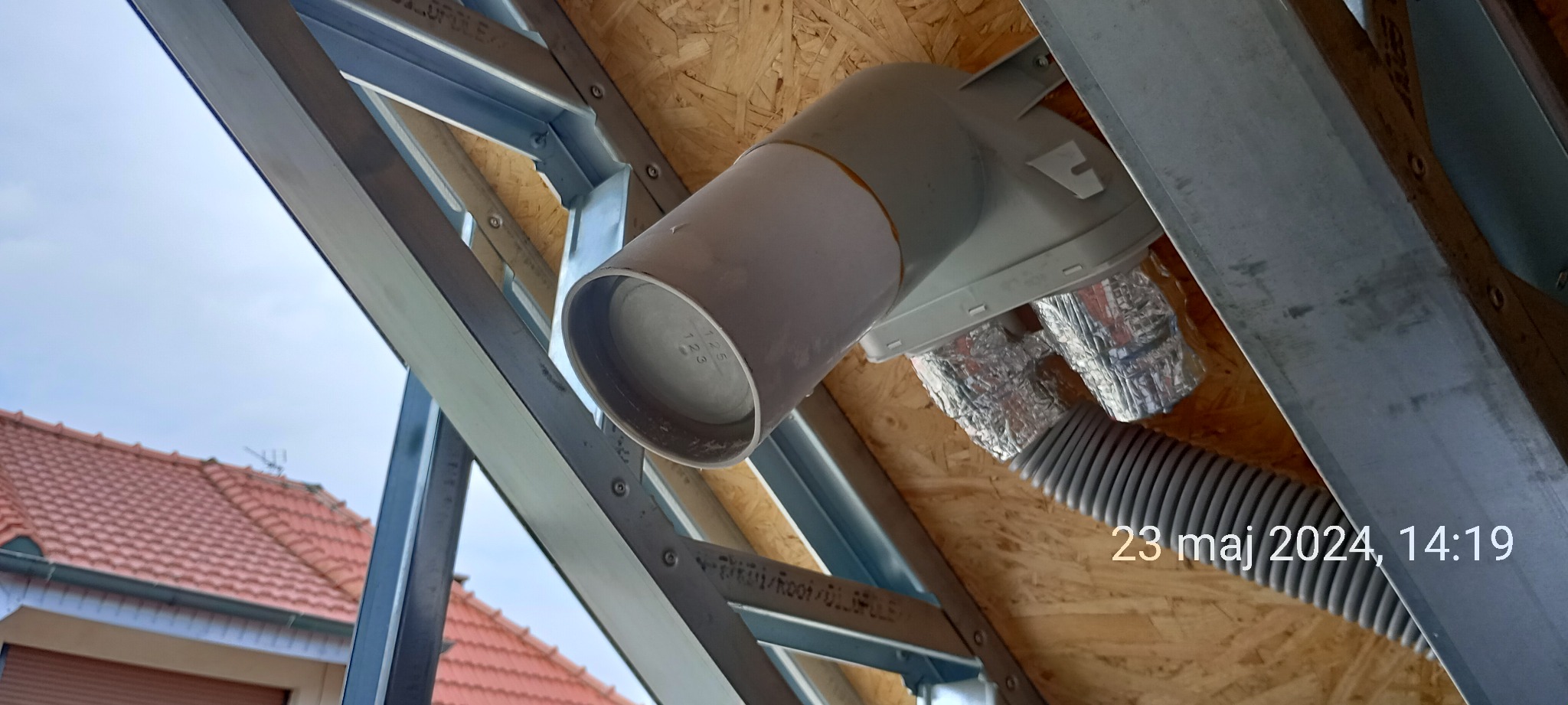 Szara rura wentylacyjna podłączona do metalowego stelażu dachu, widoczna izolacja termiczna i drewniana płyta OSB