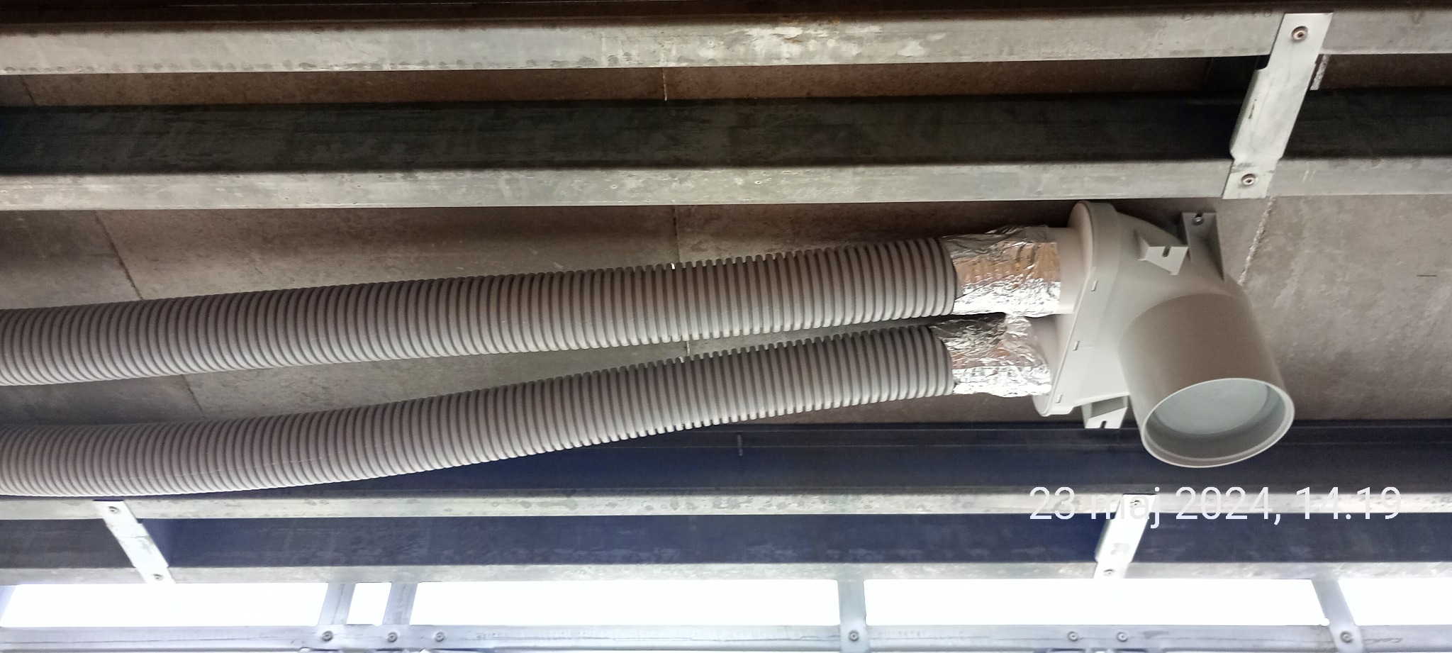 Dwa elastyczne przewody wentylacyjne przymocowane do sufitu za pomocą metalowych wsporników i połączone z białym elementem wentylacyjnym, zabezpieczone folią aluminiową.