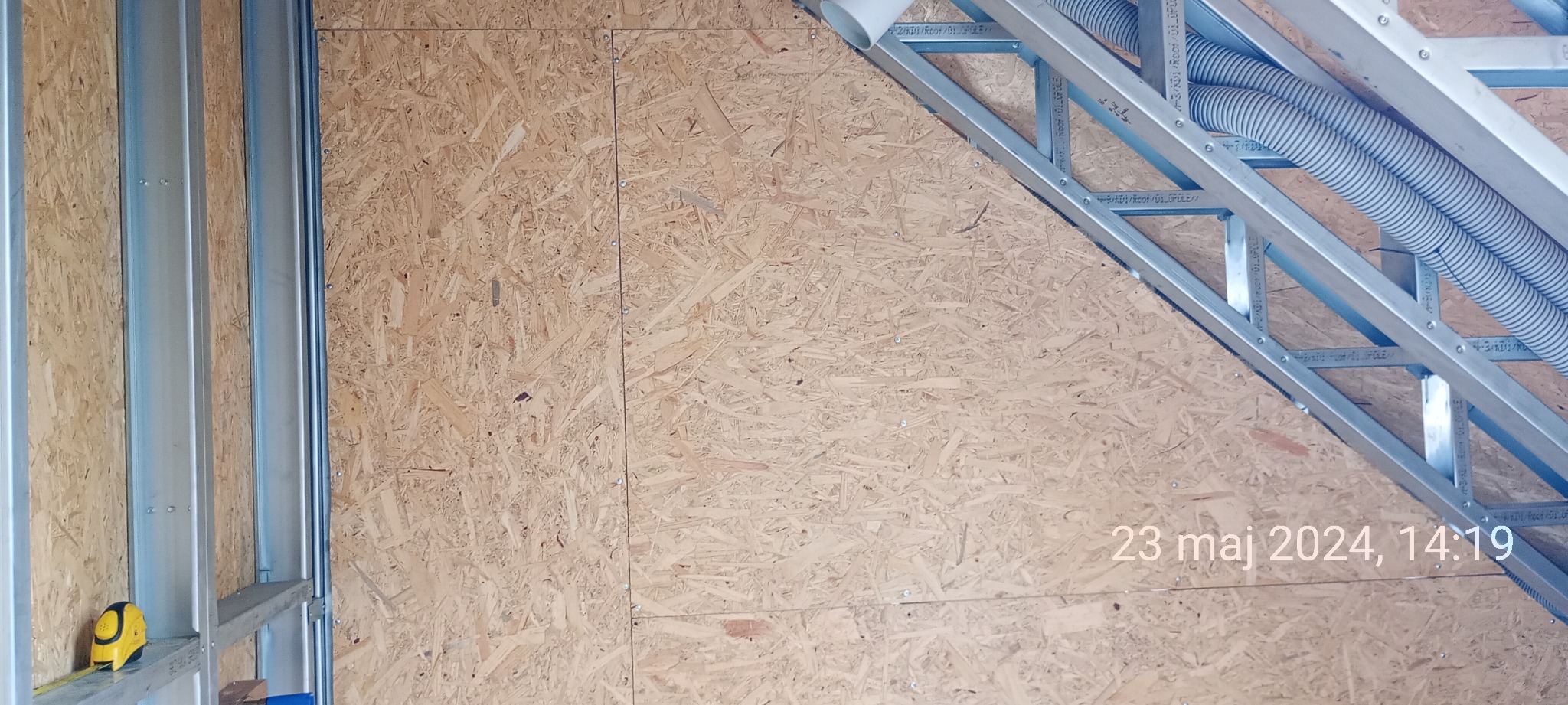 Wnętrze w trakcie budowy: ściana z płyty OSB na metalowym stelażu, widoczne elementy konstrukcji dachu i rury wentylacyjne, żółta miarka na dole po lewej stronie.