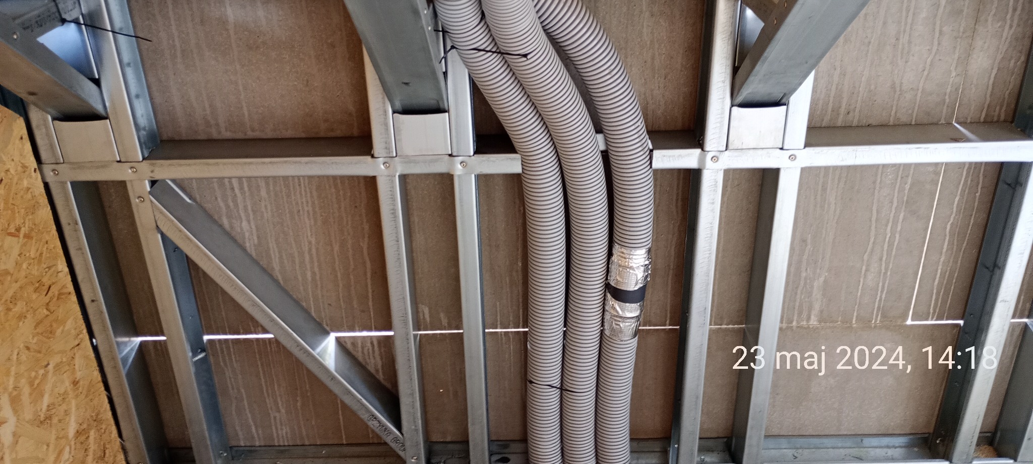 Widok od dołu na szkieletową konstrukcję sufitu z trzema elastycznymi rurami instalacyjnymi, zabezpieczonymi folią aluminiową i czarną taśmą, przymocowanymi do metalowego stelażu.