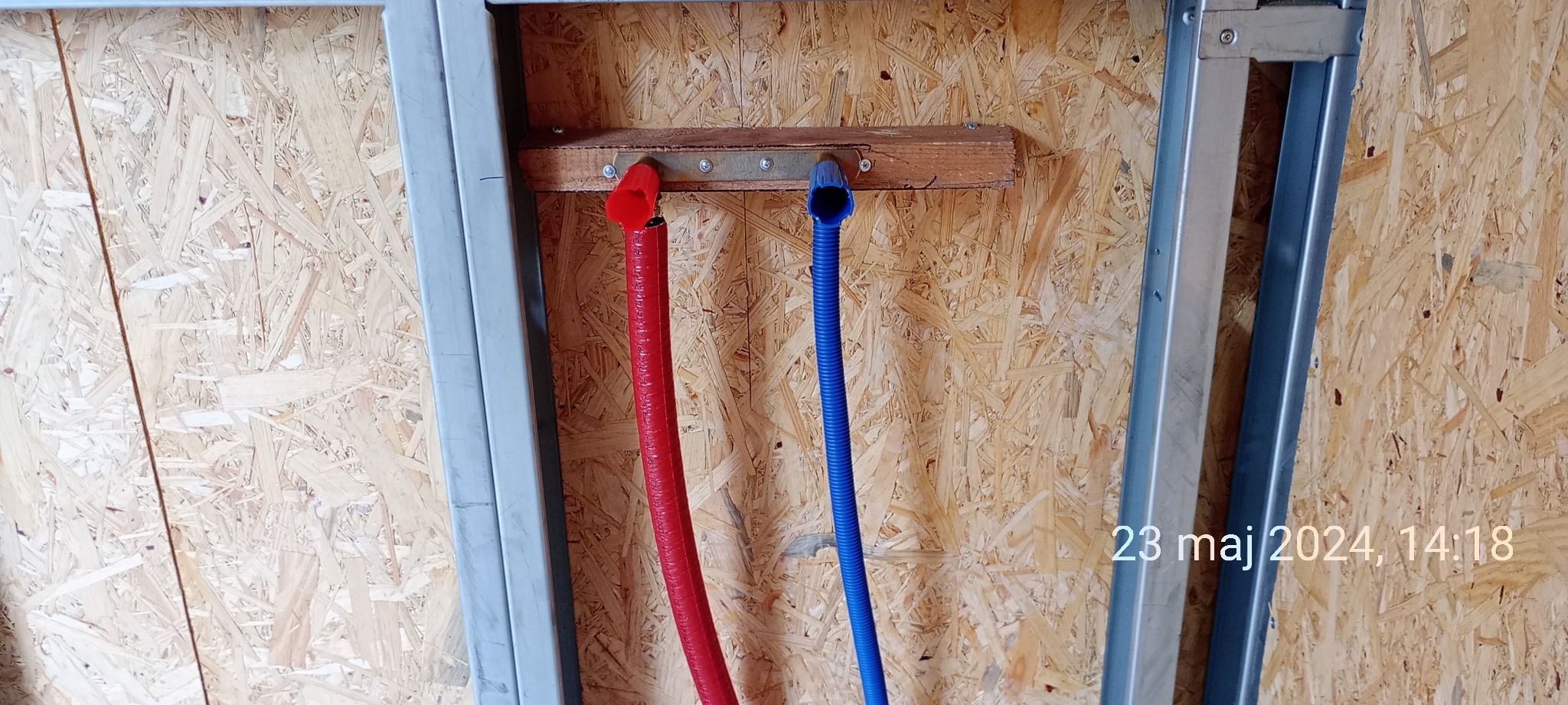 Czerwona i niebieska rura wodna przymocowane do drewnianej belki na ścianie z płyty OSB, widoczne metalowe profile konstrukcyjne.