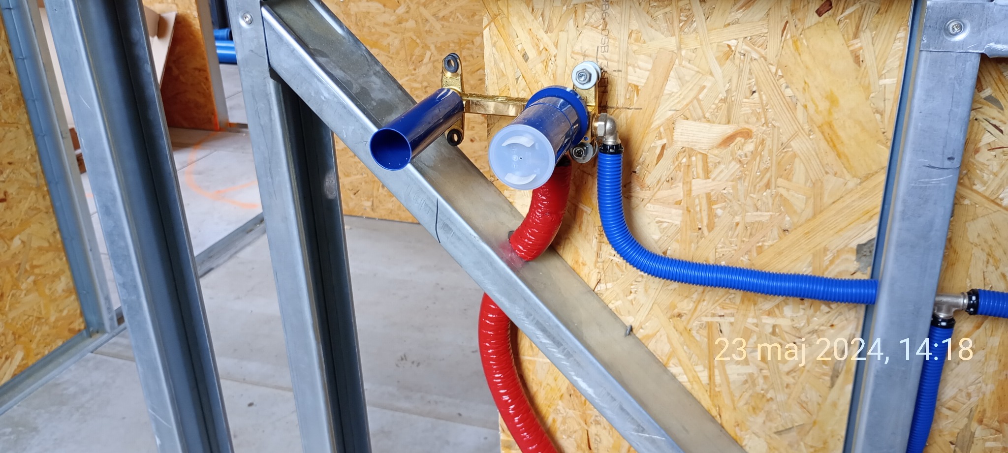 Instalacja wodna w ścianie szkieletowej: niebieskie i czerwone rury doprowadzające wodę zamontowane na metalowym stelażu, widoczne elementy mosiężne.