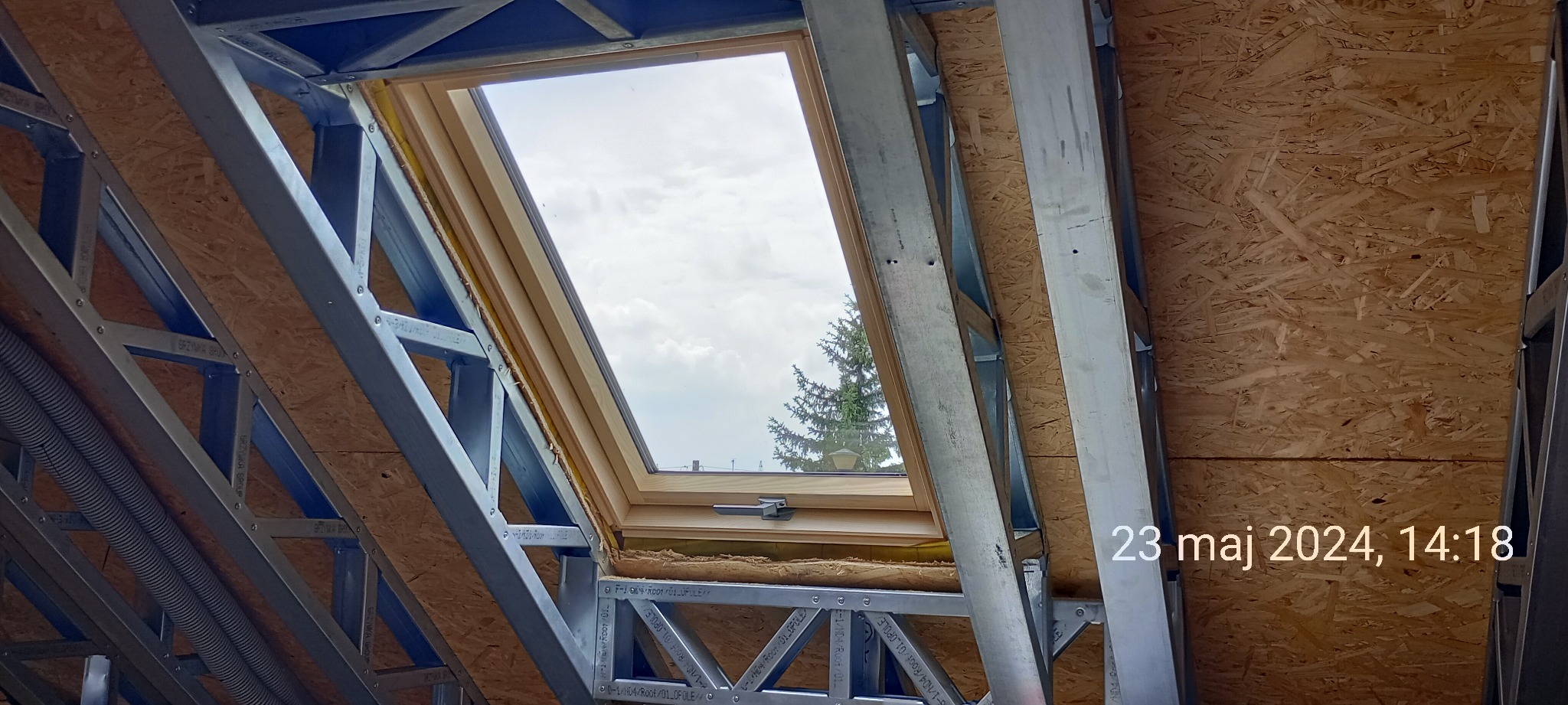 Wnętrze poddasza w trakcie budowy domu szkieletowego, widoczna konstrukcja metalowa, płyta OSB i okno dachowe z widokiem na drzewo iglaste.