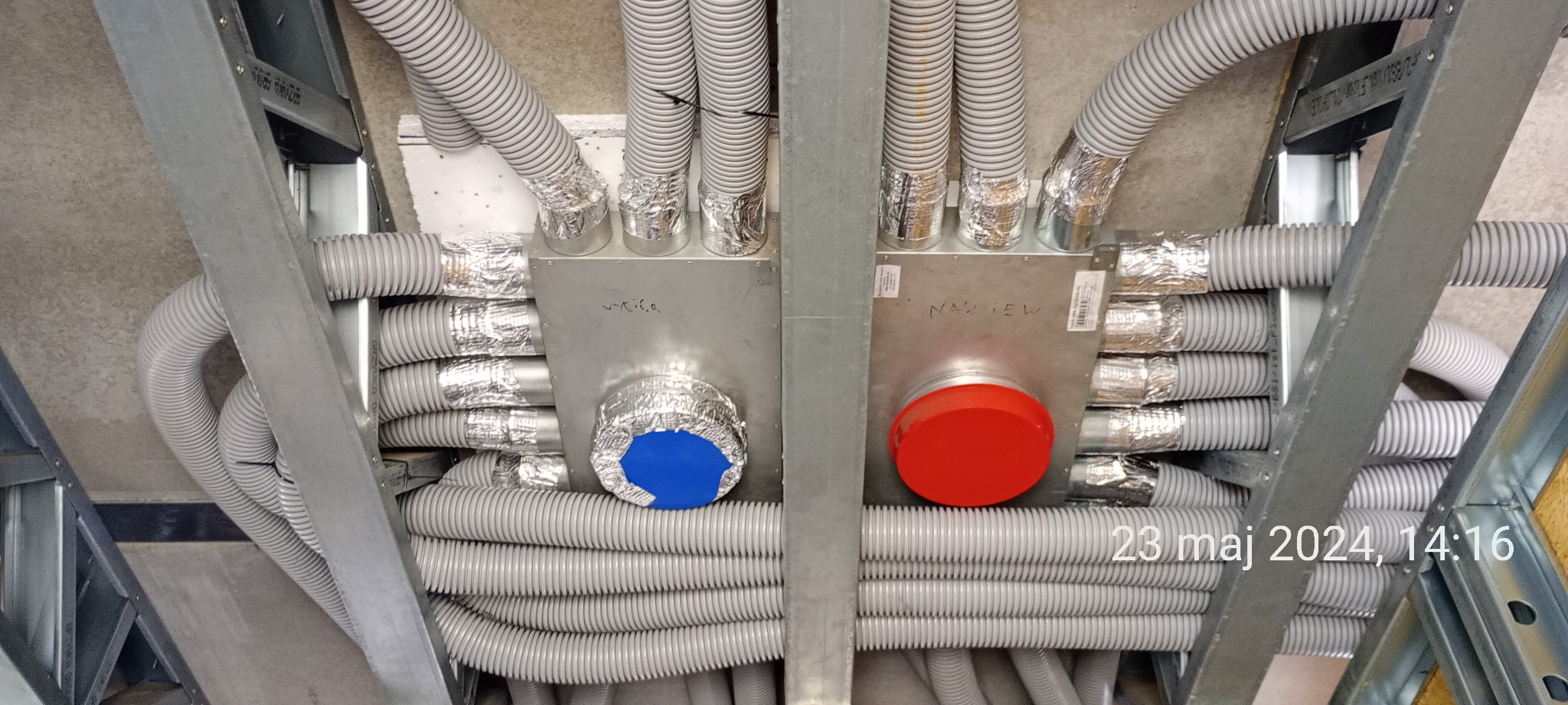 Widok instalacji wentylacyjnej z szarymi, elastycznymi przewodami, podwieszonymi pod stropem na metalowej konstrukcji, z widocznymi dwoma metalowymi skrzynkami z czerwonym i niebieskim okrągłym...