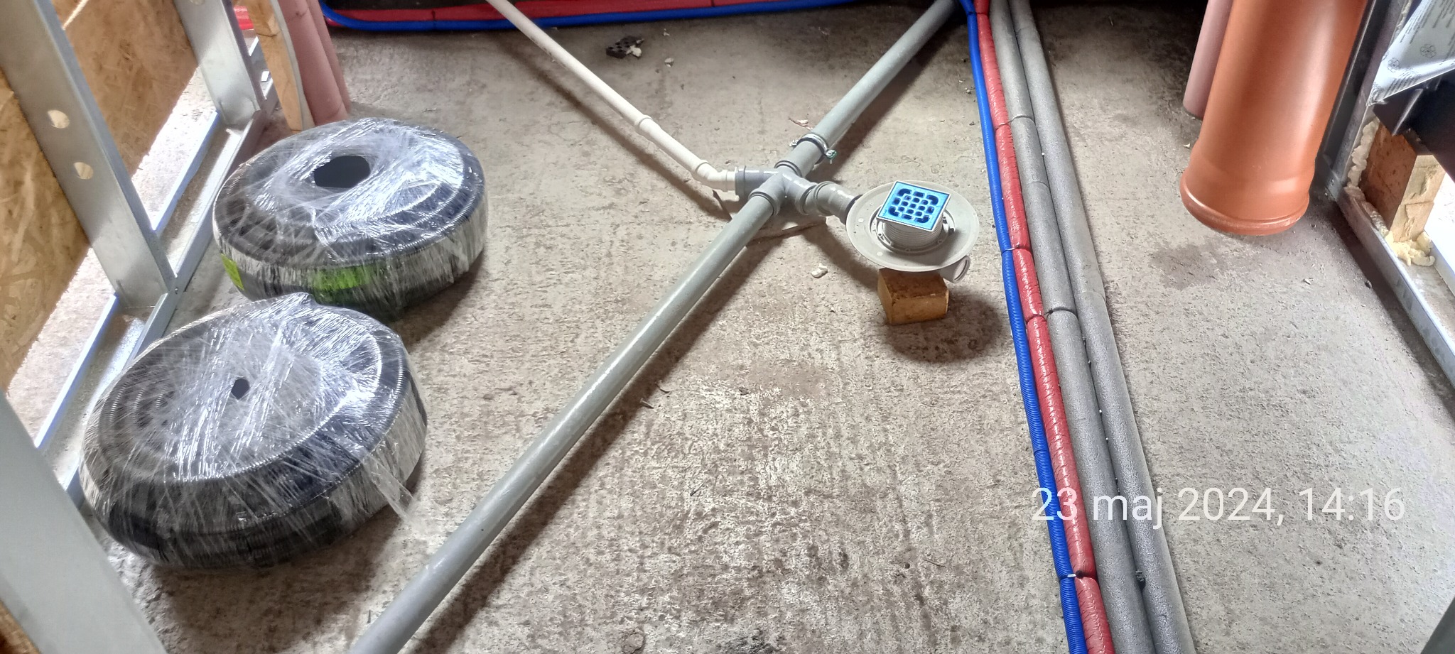 Instalacja kanalizacyjna i rury grzewcze w trakcie montażu w surowym stanie, z widocznymi rolkami rur w folii i datą na zdjęciu.