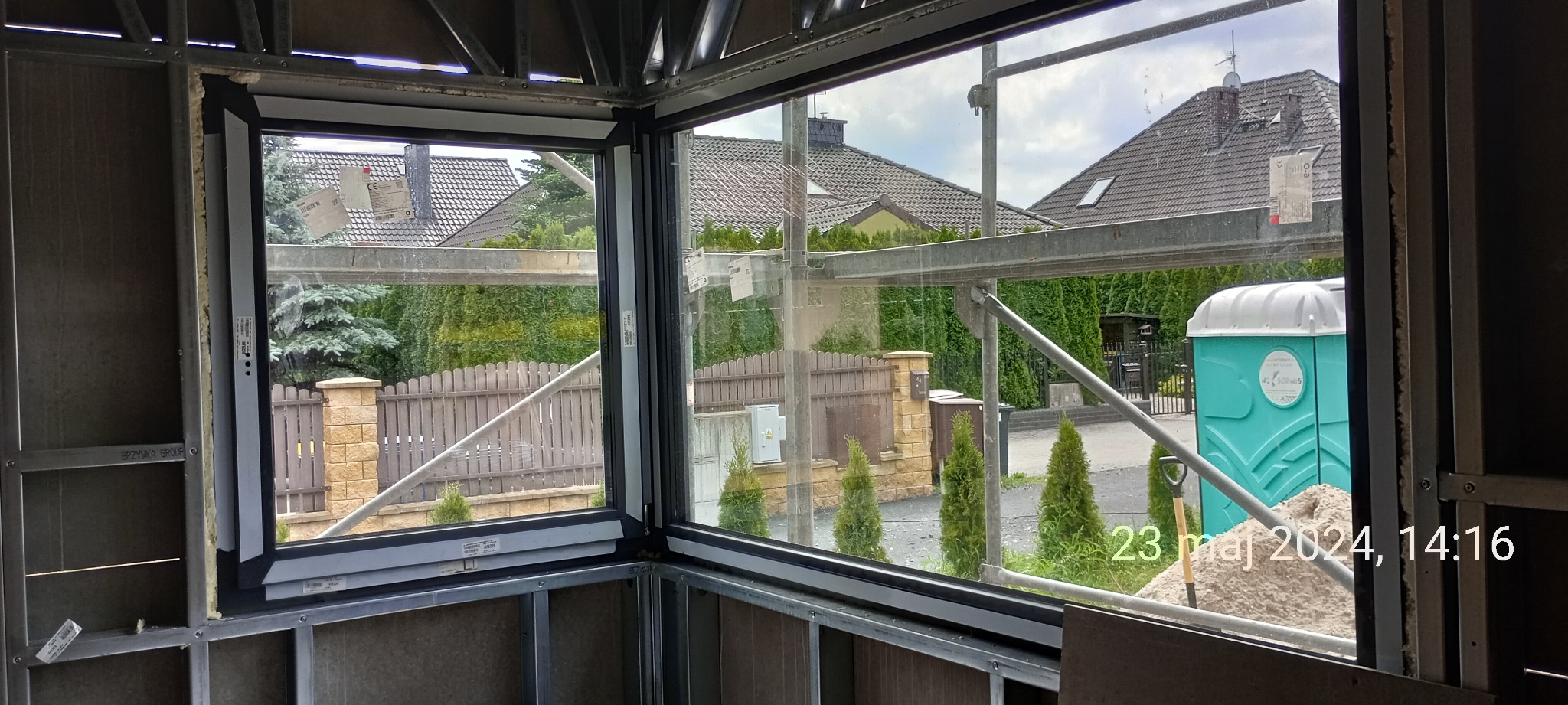 Surowy stan domu szkieletowego w Warszawie: widok przez okno na ogrodzenie, zieleń i przenośną toaletę na zewnątrz.