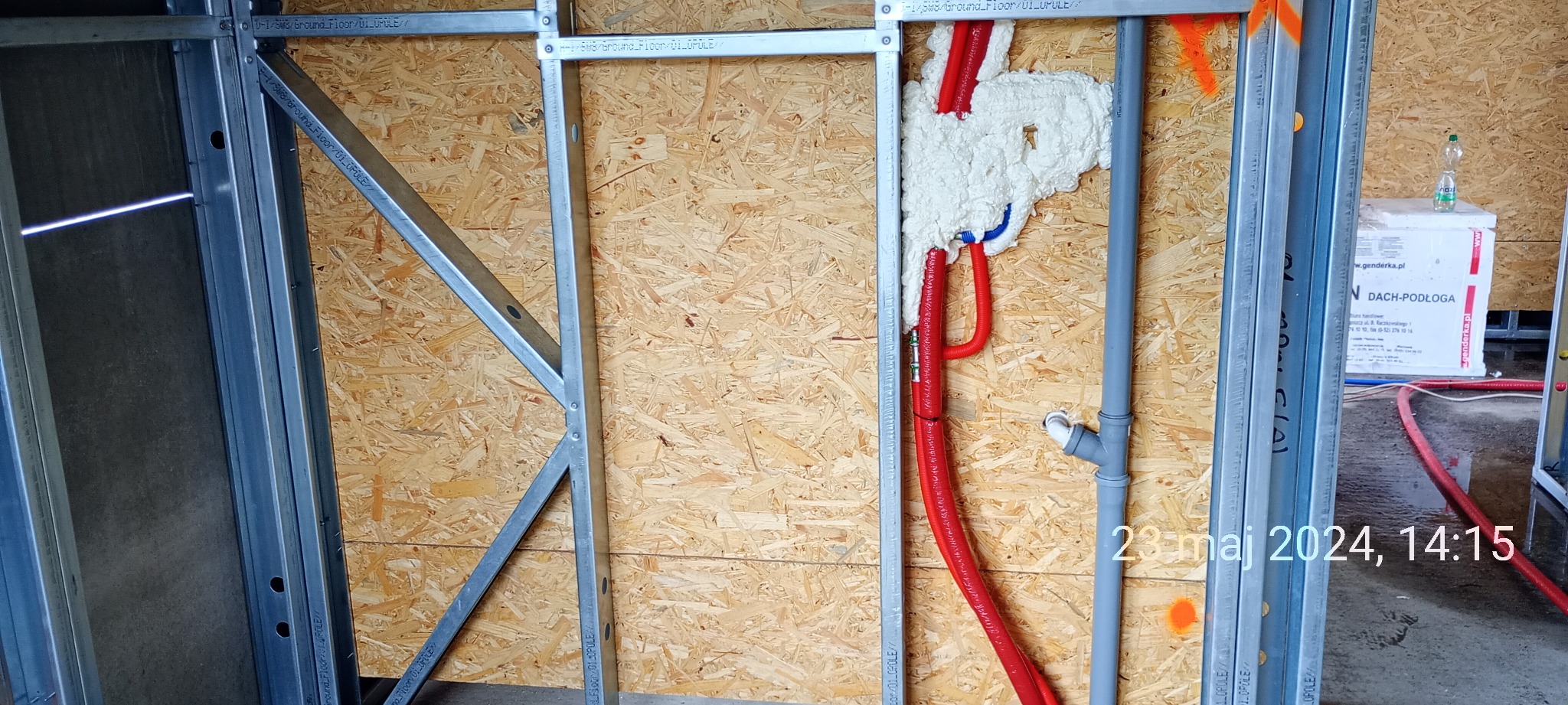 Konstrukcja domu szkieletowego w trakcie budowy: widoczne metalowe profile, płyta OSB, izolacja piankowa wokół rur instalacyjnych oraz szare rury kanalizacyjne.