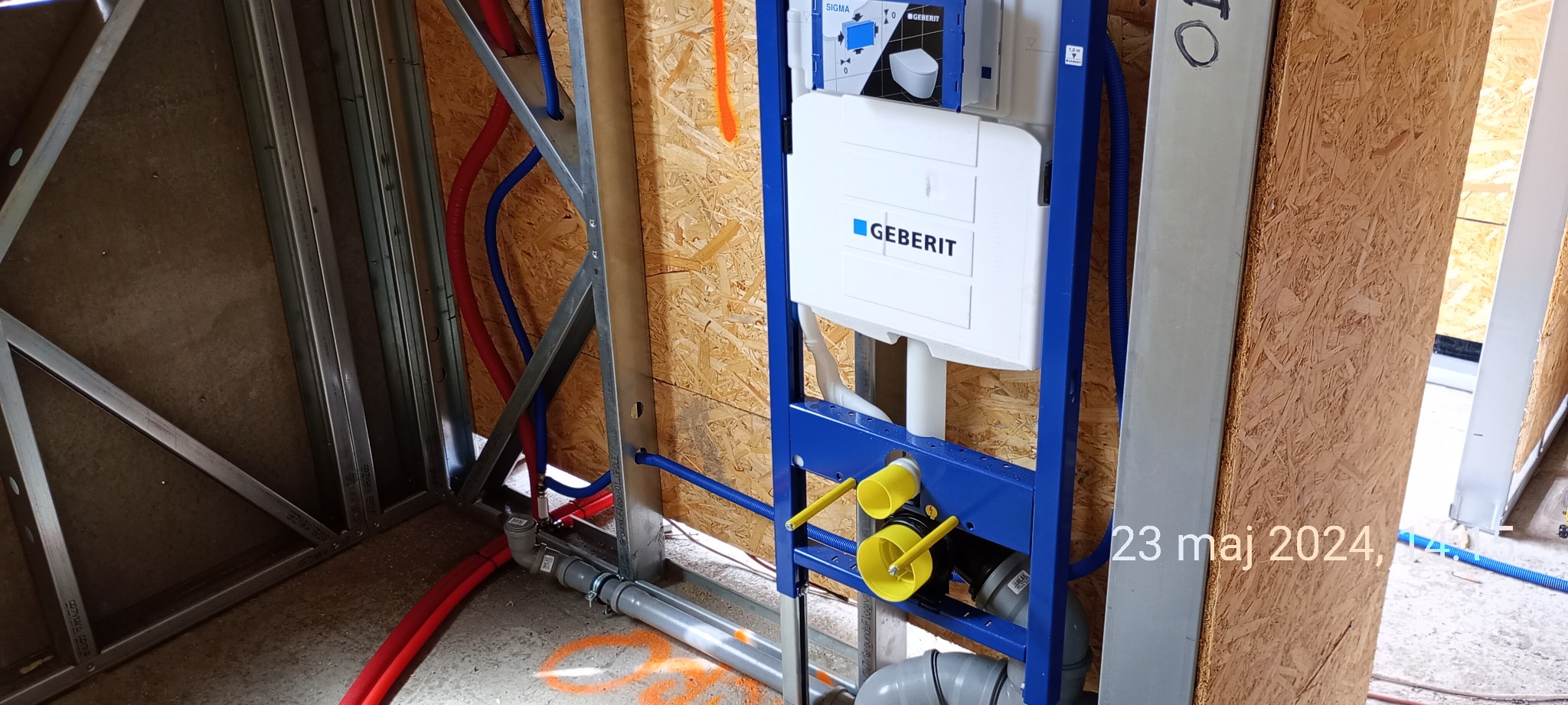 Stelaż podtynkowy Geberit zamontowany w konstrukcji szkieletowej z widocznymi rurami doprowadzającymi wodę i odpływowymi, przed zabudową płytami gipsowo-kartonowymi.