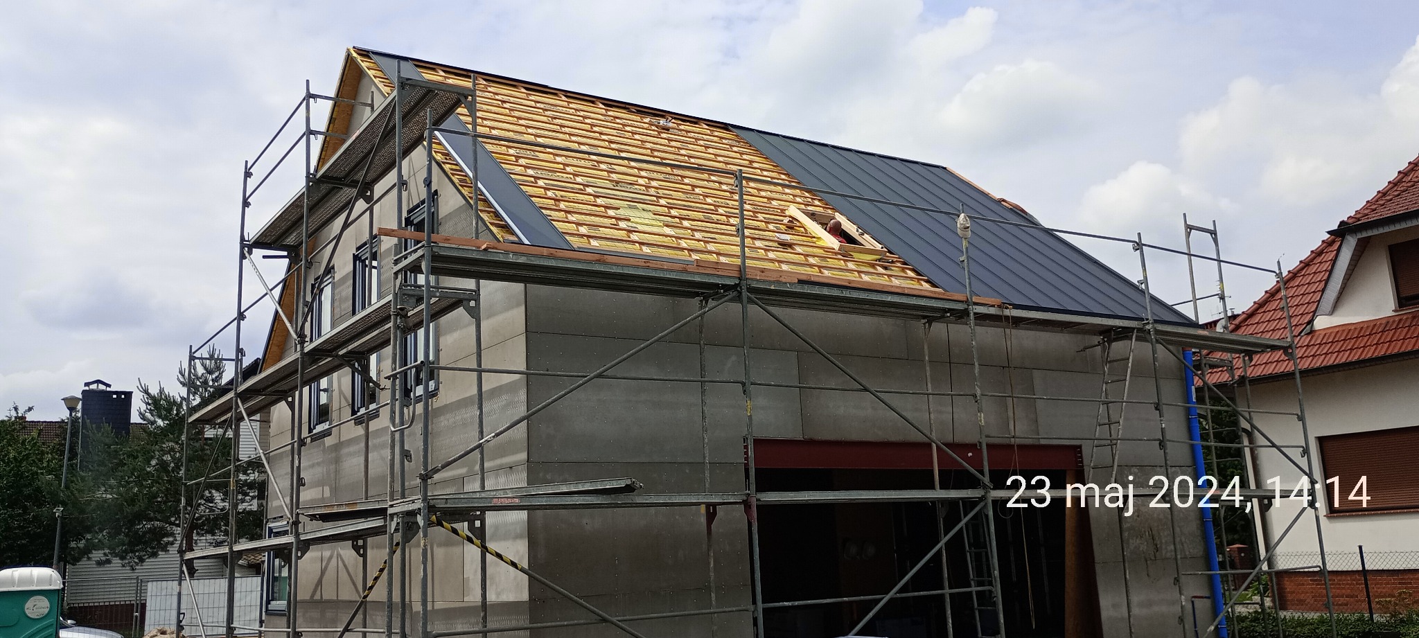Dom w trakcie budowy: widoczne rusztowanie, ściany pokryte szarymi panelami, częściowo ułożona dachówka w kolorze grafitowym oraz drewniana konstrukcja dachu z oknem dachowym, na którym pracuje osoba.