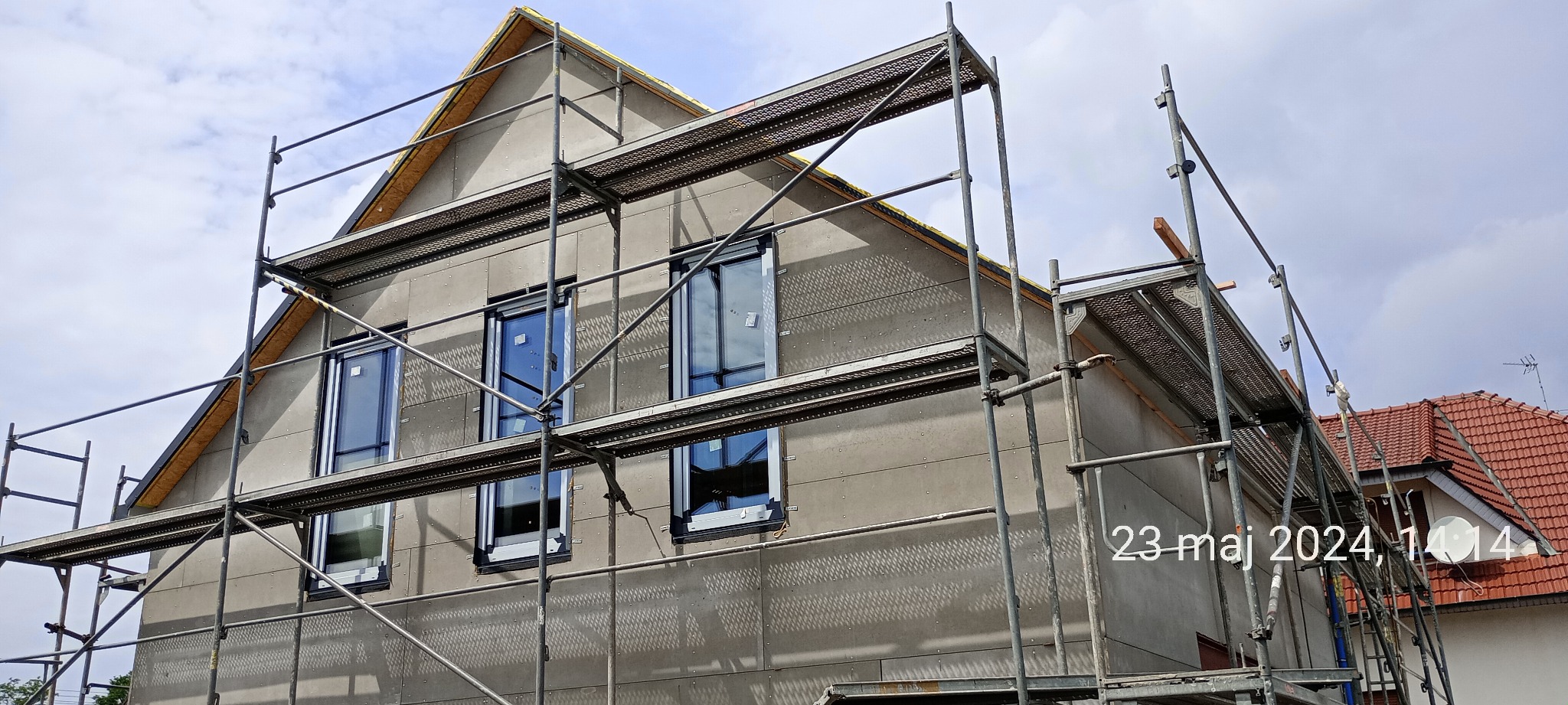 Surowy dom w trakcie budowy z widocznymi rusztowaniami i oknami, z zewnętrzną warstwą szarej płyty izolacyjnej z charakterystycznym wzorem perforacji, widok na sąsiedni dom z czerwoną dachówką.