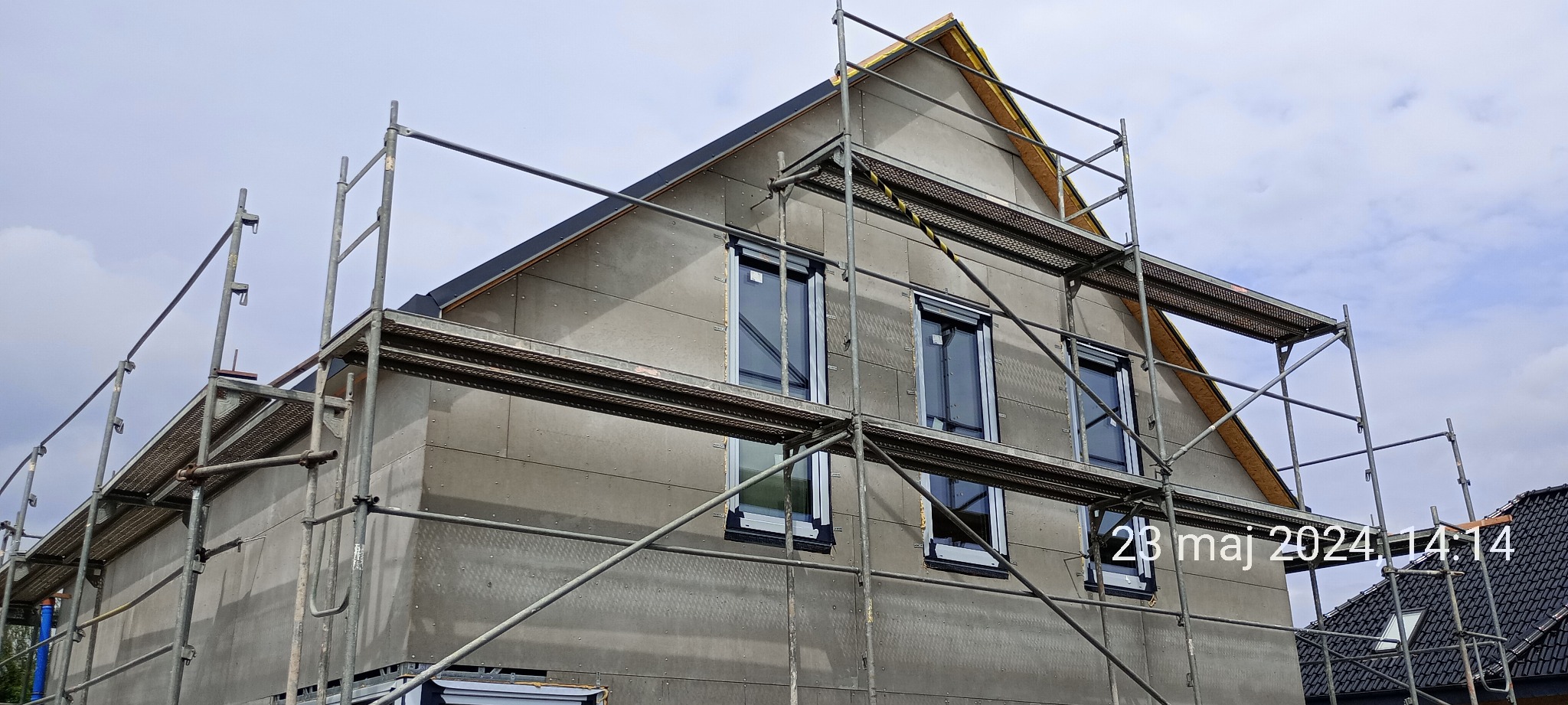 Surowa elewacja domu w trakcie budowy, widoczne rusztowania, okna z roletami zewnętrznymi, szara struktura elewacji z widocznymi mocowaniami.