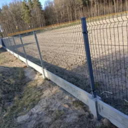 Nowoczesne ogrodzenie z paneli na betonowej podmurówce, widok z boku na tle nagiego lasu w słoneczny dzień, z widocznym fragmentem białego samochodu dostawczego po lewej stronie.