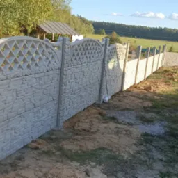 Długi, dekoracyjny, betonowy płot z elementami imitującymi kamień i ażurową górną częścią, ciągnący się wzdłuż działki, z widocznym samochodem dostawczym w tle.