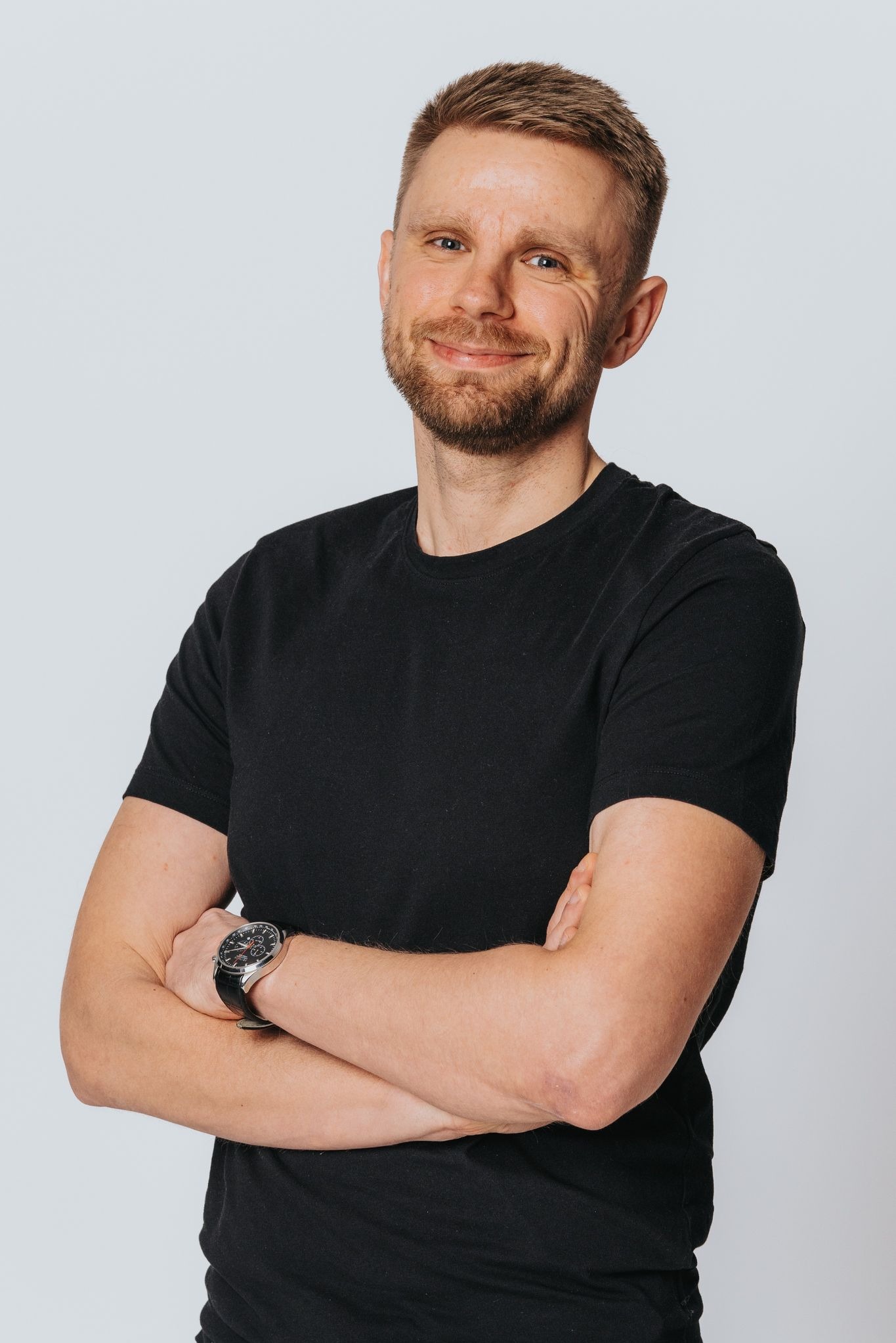 Portret uśmiechniętego mężczyzny z brodą, w czarnym T-shircie, z założonymi rękami i zegarkiem na nadgarstku, na jasnym tle.
