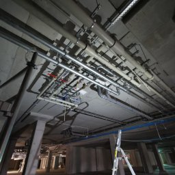 Oleksandr Misiiuk - Skomplikowany system rur i przewodów pod betonowym stropem w surowym stanie, widoczna drabina aluminiowa i fragment otwartej przestrzeni.