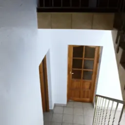 Widok z góry na klatkę schodową z drewnianymi schodami i balustradą, białe ściany, dwoje drewnianych drzwi z przeszkleniami i szarą podłogą z płytek.