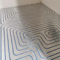 Rozłożona instalacja ogrzewania podłogowego z niebieskimi rurkami na srebrnej folii izolacyjnej, widok z góry w surowym, nieotynkowanym pomieszczeniu.