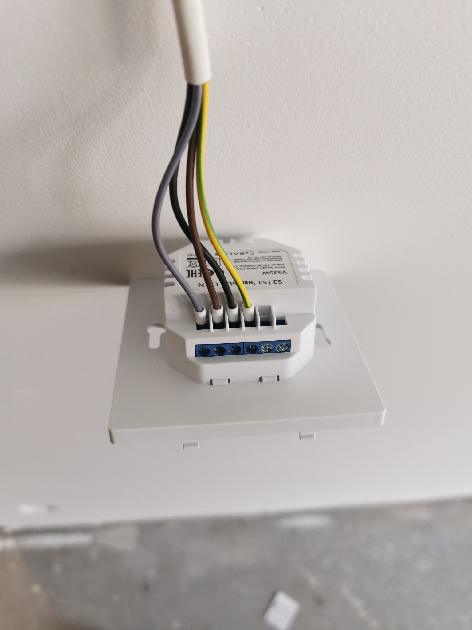 Podłączenie przewodów elektrycznych do białego modułu sterującego, widoczne kolorowe kable (szary, brązowy, żółto-zielony) wpięte w złącza śrubowe.