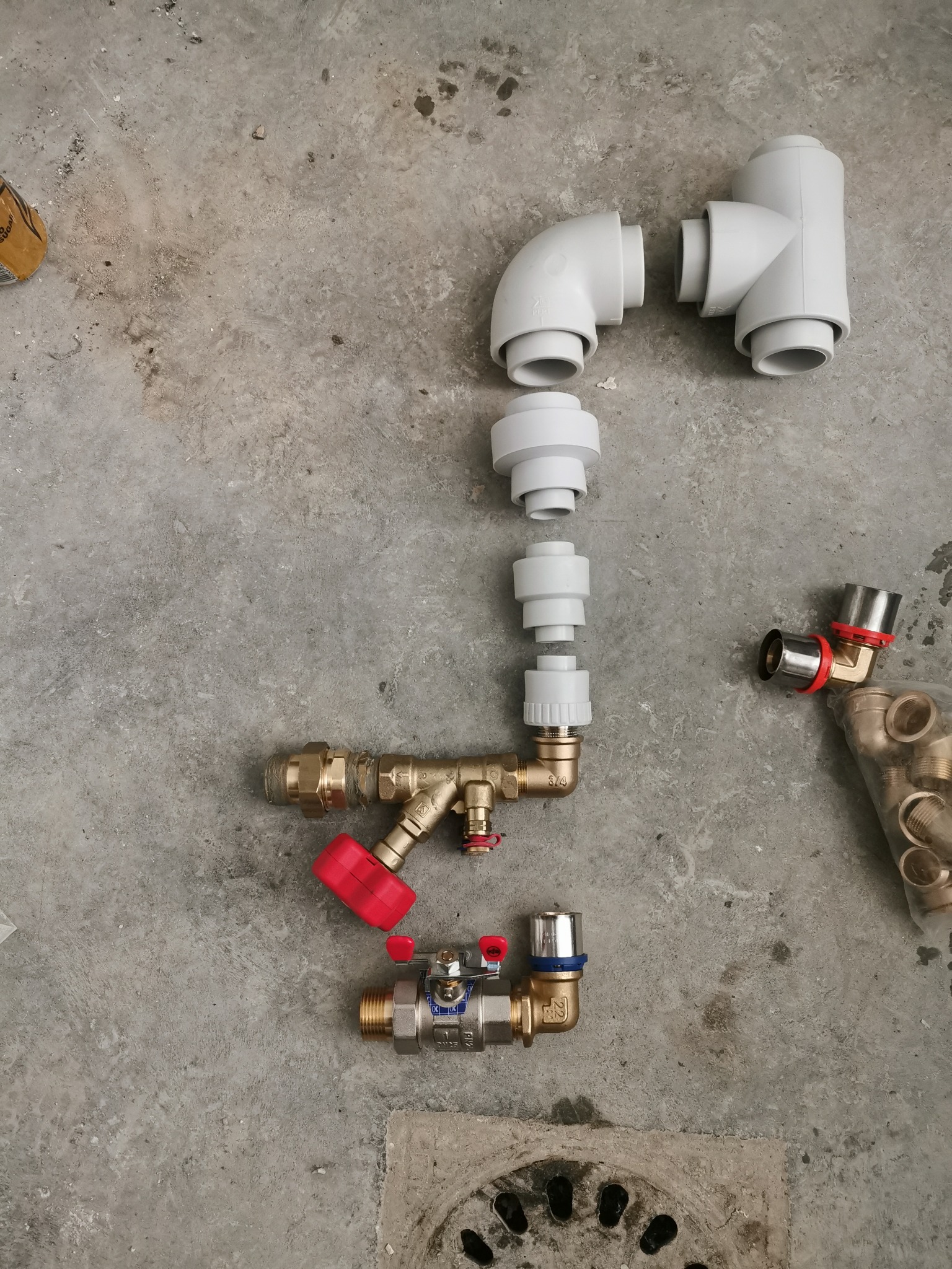 Elementy instalacji hydraulicznej złączone ze sobą na szarym, betonowym podłożu. Widoczne kształtki z tworzywa sztucznego, zawory z czerwonymi pokrętłami i metalowe kolanka. Część elementów jeszcze...