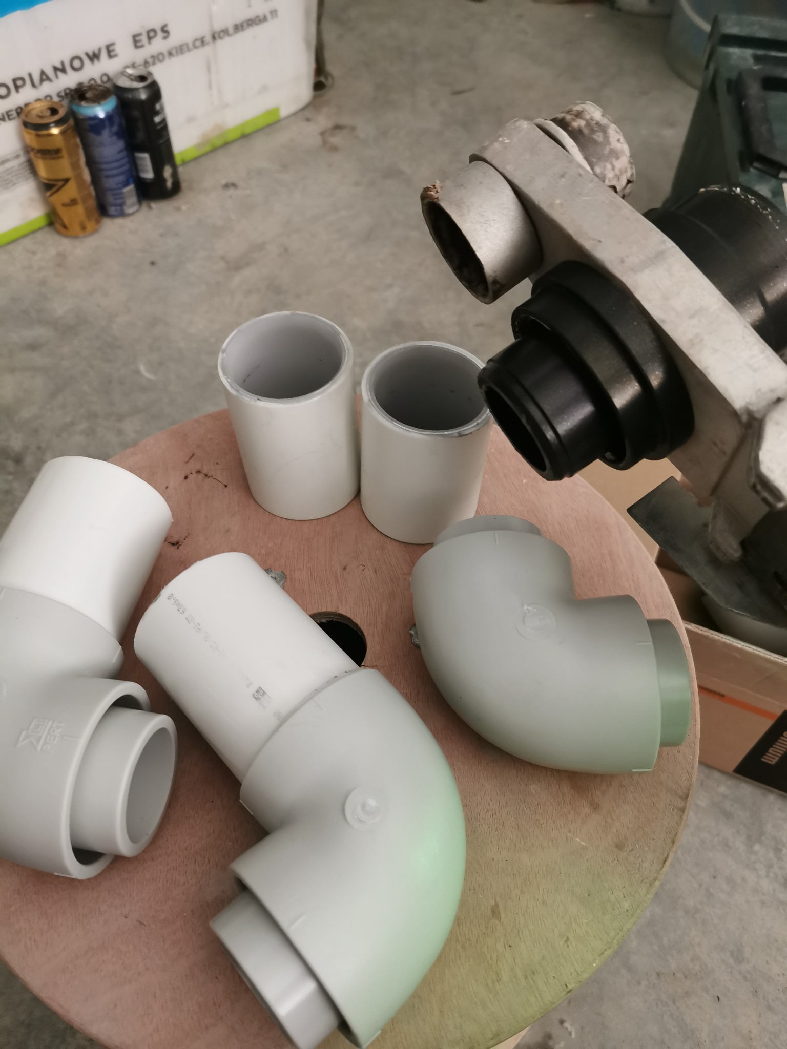 Przygotowane elementy instalacji hydraulicznej: białe rury, kolanka i złączki PVC leżące na okrągłym drewnianym blacie obok narzędzia do cięcia rur. W tle widoczne puszki po napojach i karton.