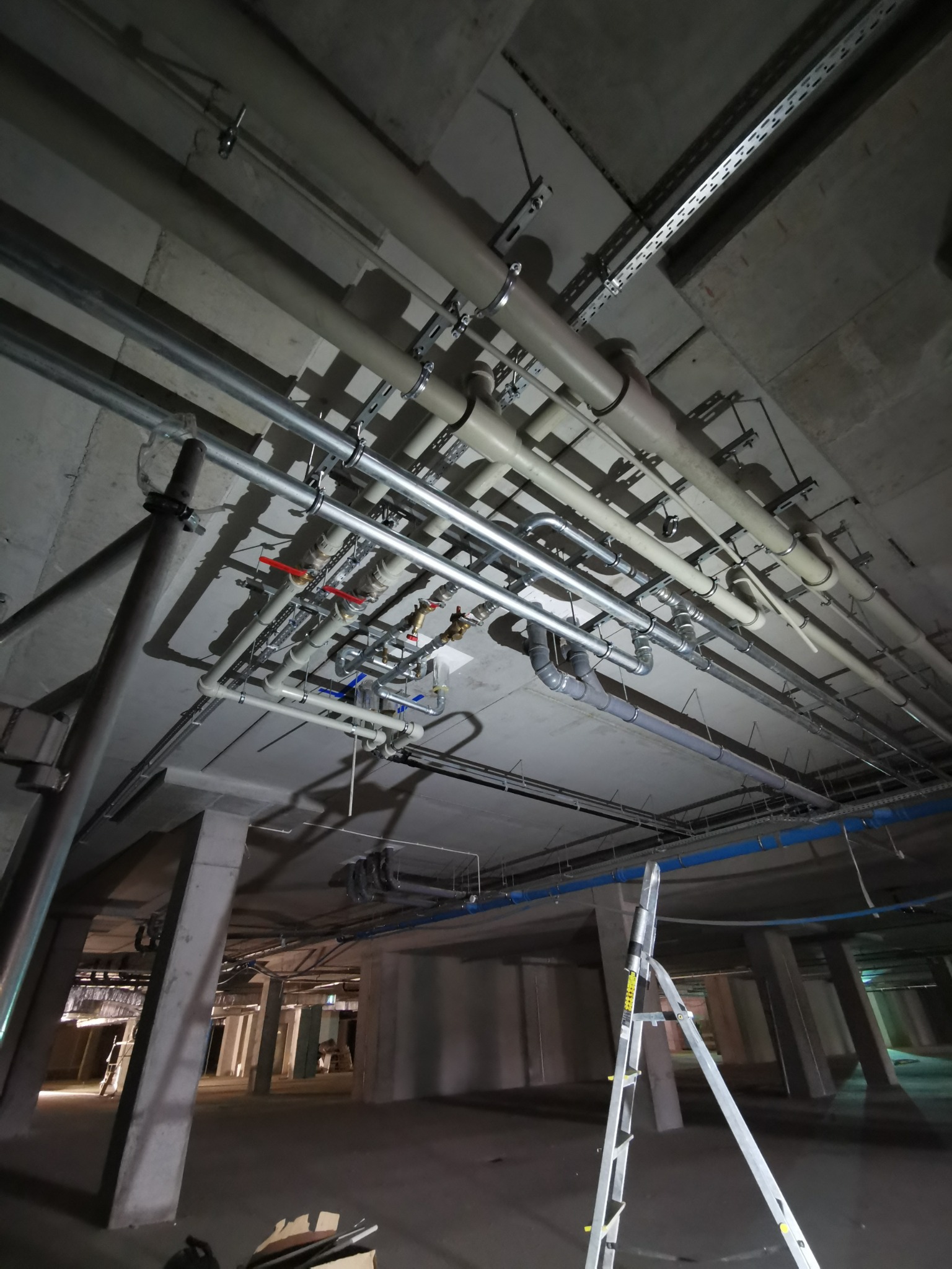 Skomplikowany system rur i przewodów pod betonowym stropem w surowym stanie, widoczna drabina aluminiowa i fragment otwartej przestrzeni.