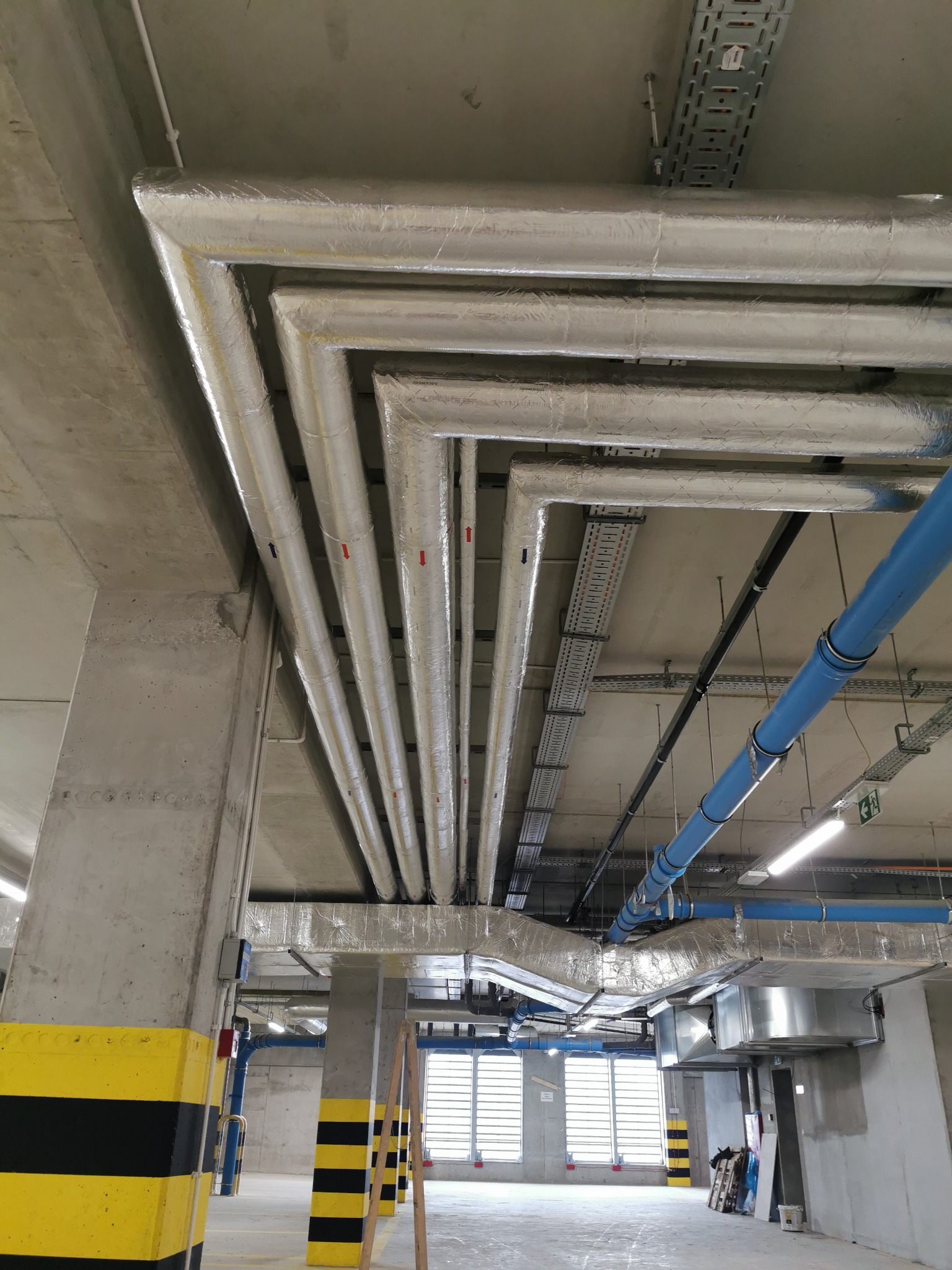 Widok instalacji rur w izolacji termicznej pod sufitem parkingu podziemnego, widoczne elementy wentylacji, rury niebieskie, koryta kablowe i słupy z oznaczeniami ostrzegawczymi.