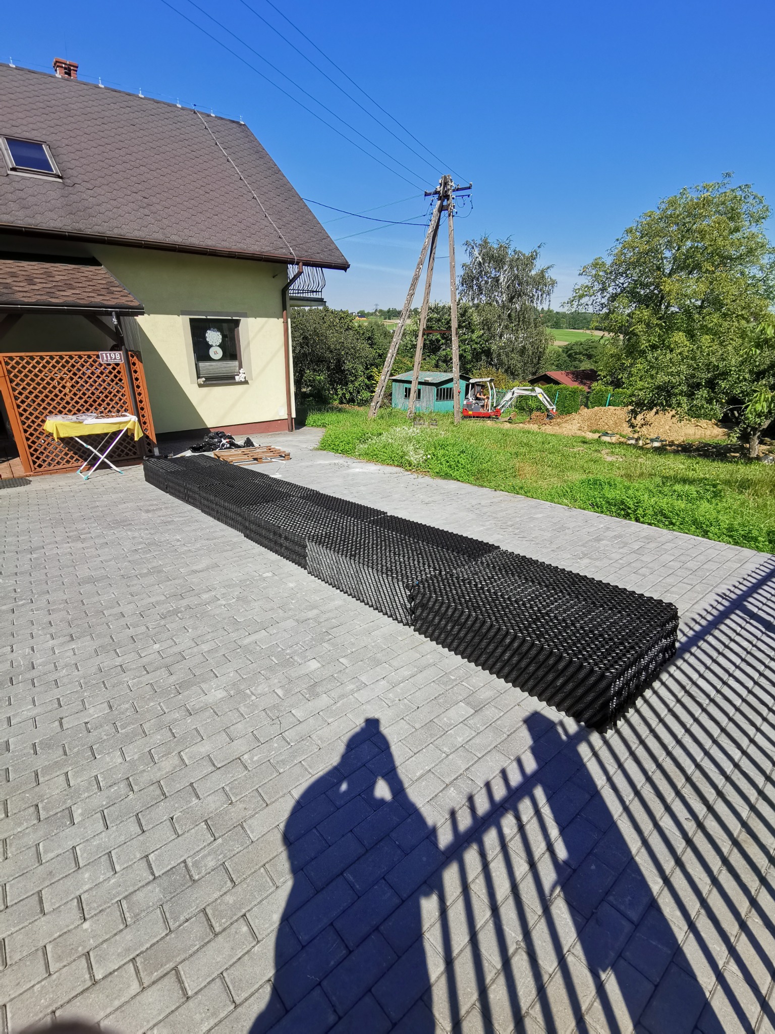 Czarne moduły rozsączające ułożone na szarym bruku przed domem z ogrodem, w tle minikoparka podczas prac ziemnych.