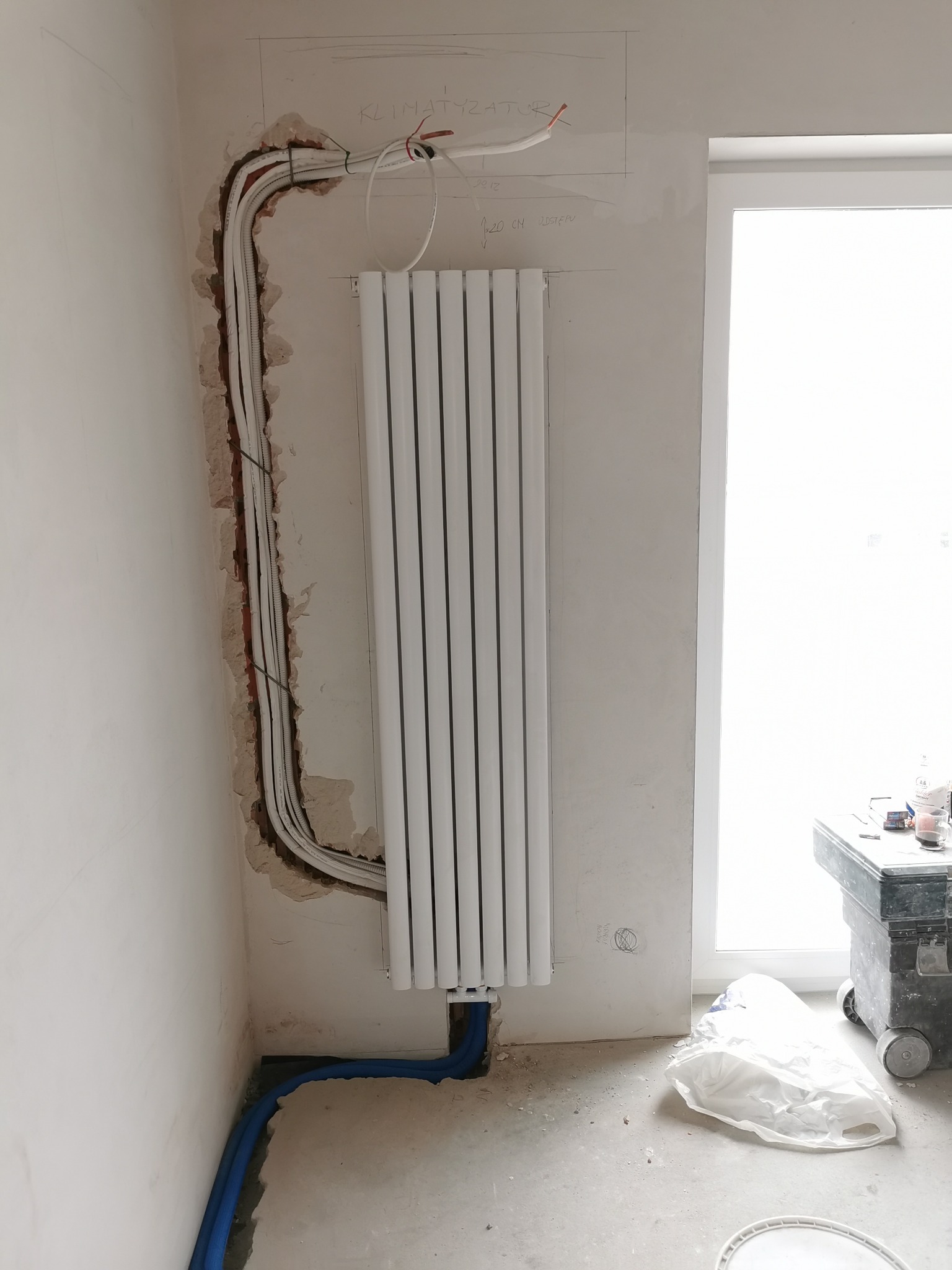 Biały grzejnik panelowy zamontowany na ścianie, z widocznymi niebieskimi rurami doprowadzającymi wodę i wyżłobieniami w ścianie na kable elektryczne i rury klimatyzacji, w trakcie remontu.