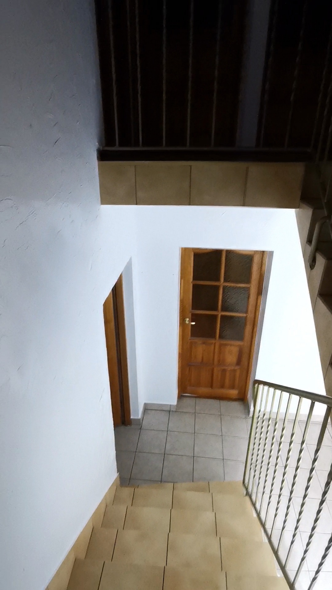 Widok z góry na klatkę schodową z drewnianymi schodami i balustradą, białe ściany, dwoje drewnianych drzwi z przeszkleniami i szarą podłogą z płytek.