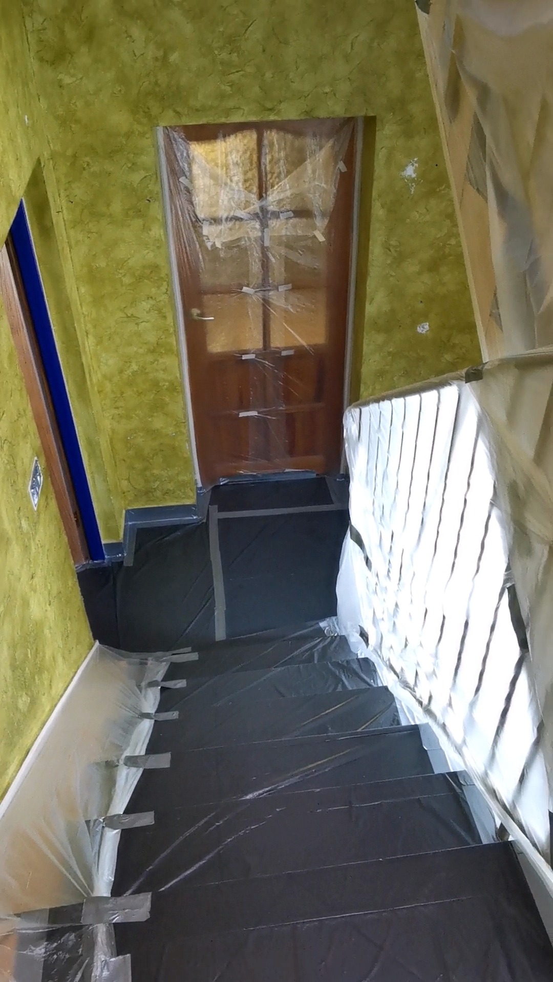 Schody i ściana zabezpieczone folią malarską przed malowaniem, widoczne naklejone taśmy ochronne, drzwi również zakryte folią.