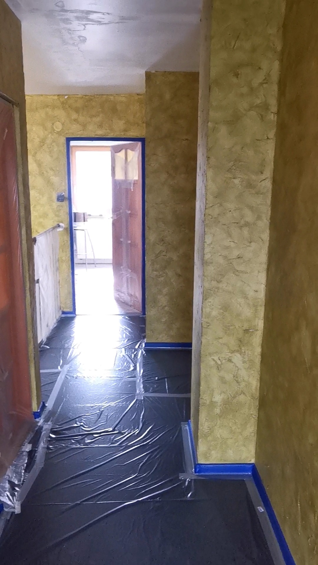 Korytarz w trakcie malowania ścian na odcienie beżu, podłoga zabezpieczona folią, framugi oklejone taśmą malarską.