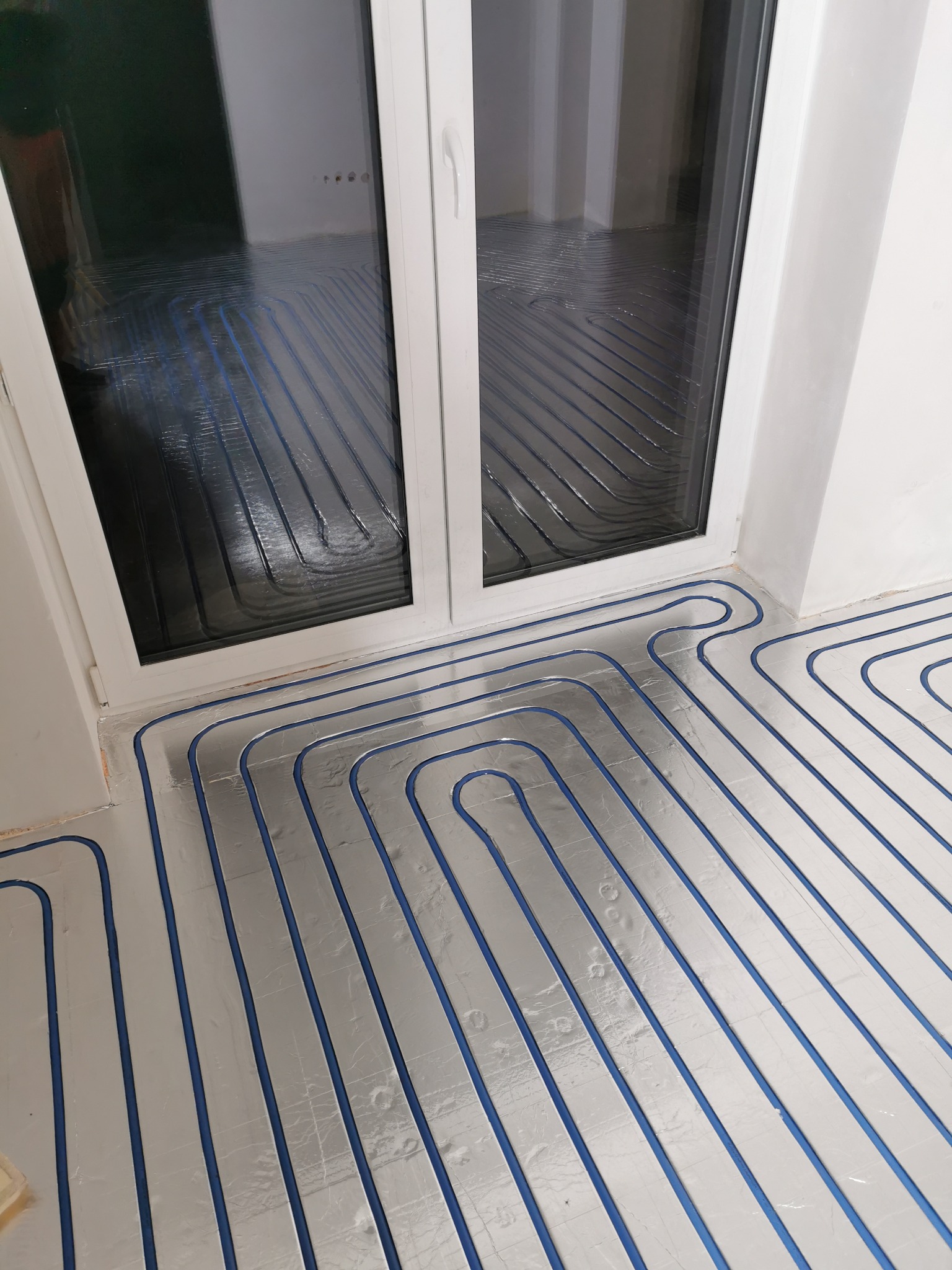 Montaż ogrzewania podłogowego: rury grzewcze w kolorze niebieskim ułożone na srebrnej folii izolacyjnej, widoczne przy oknie balkonowym.