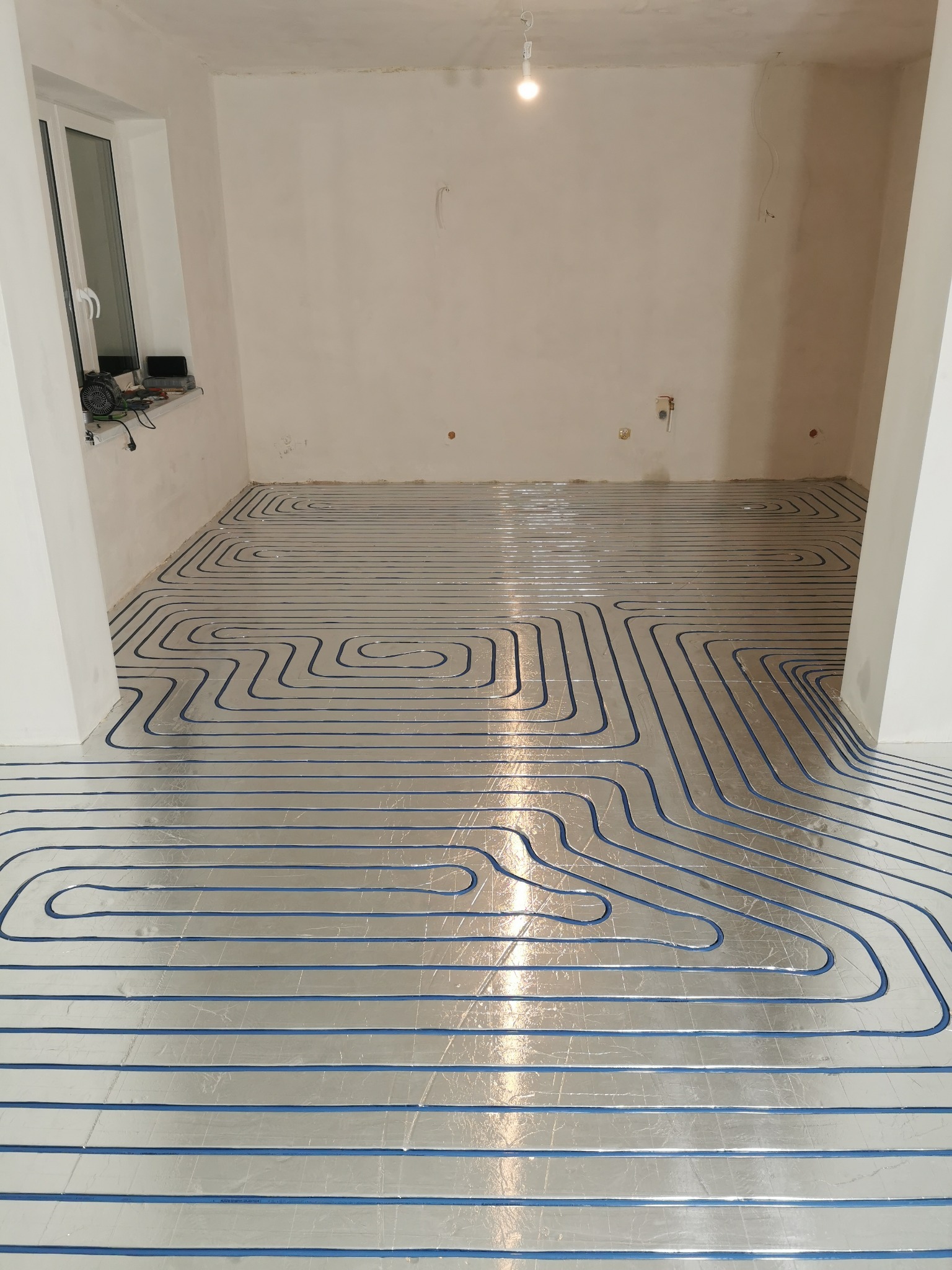 Niebieskie rury ogrzewania podłogowego ułożone na srebrnej folii izolacyjnej w jasnym, surowym wnętrzu z oknem i widocznymi śladami po instalacjach na ścianach.