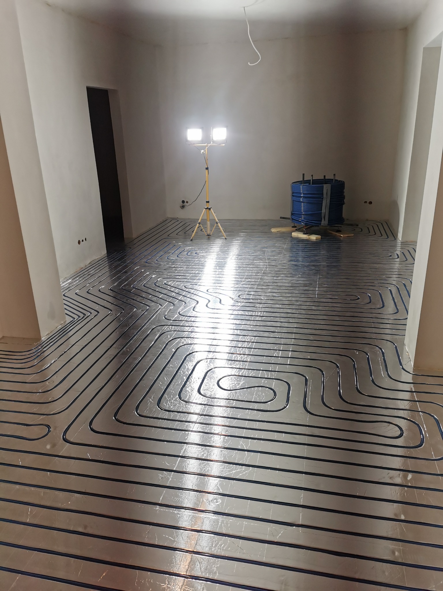 Instalacja ogrzewania podłogowego w surowym wnętrzu, widoczne rury grzewcze ułożone na srebrnej folii izolacyjnej, oświetlone lampą budowlaną, w tle zwój rur i przygotowane materiały.