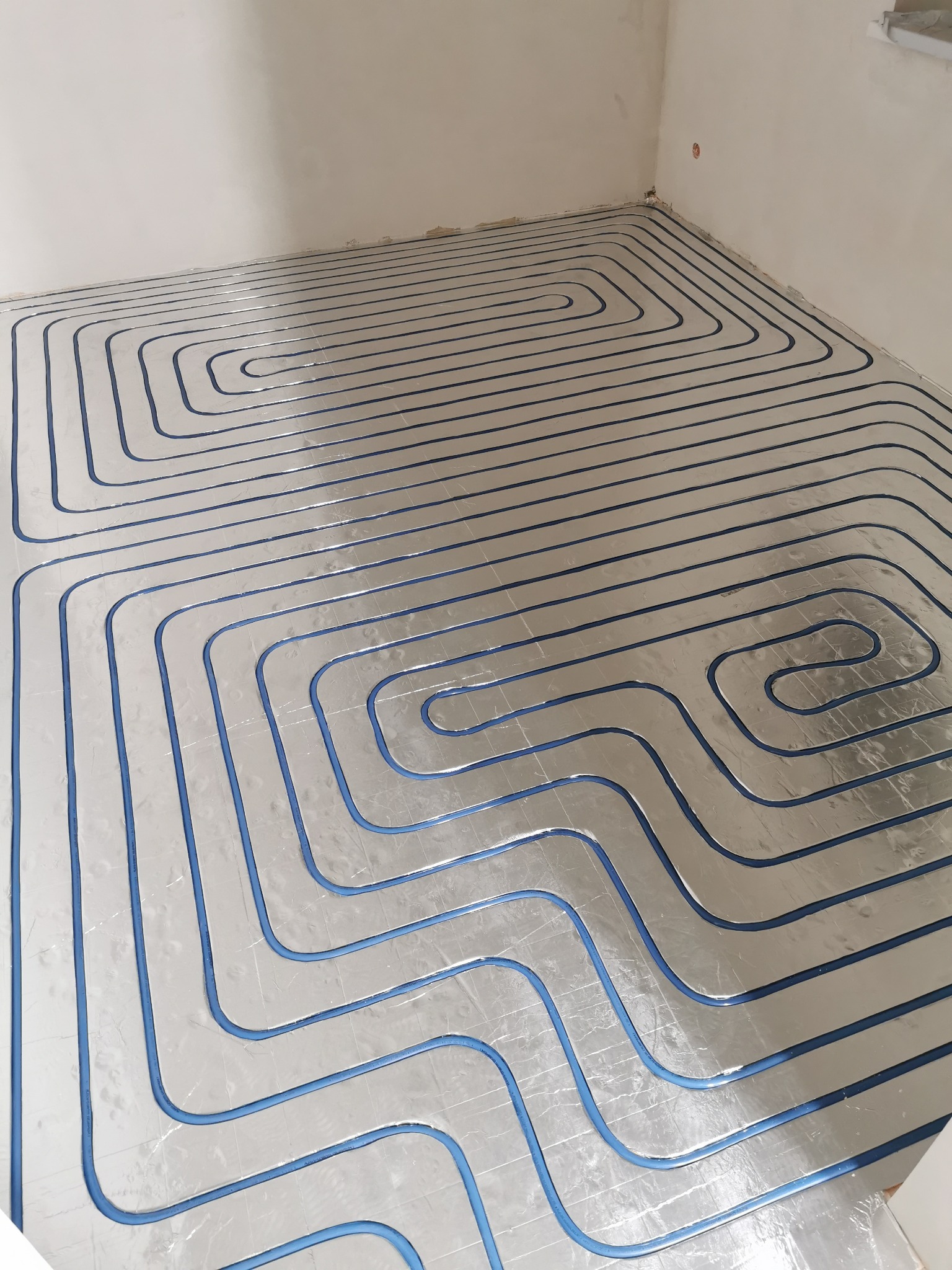 Rozłożona instalacja ogrzewania podłogowego z niebieskimi rurkami na srebrnej folii izolacyjnej, widok z góry w surowym, nieotynkowanym pomieszczeniu.