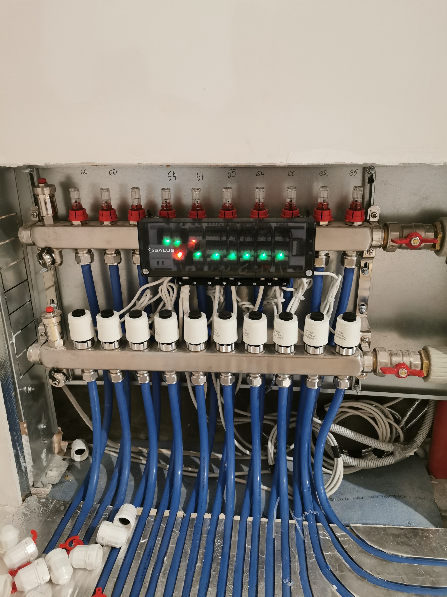 Rozdzielacz ogrzewania podłogowego z podłączonymi niebieskimi rurami, widoczne zawory termostatyczne z białymi głowicami i elektroniczny sterownik Salus z zielonymi diodami LED.