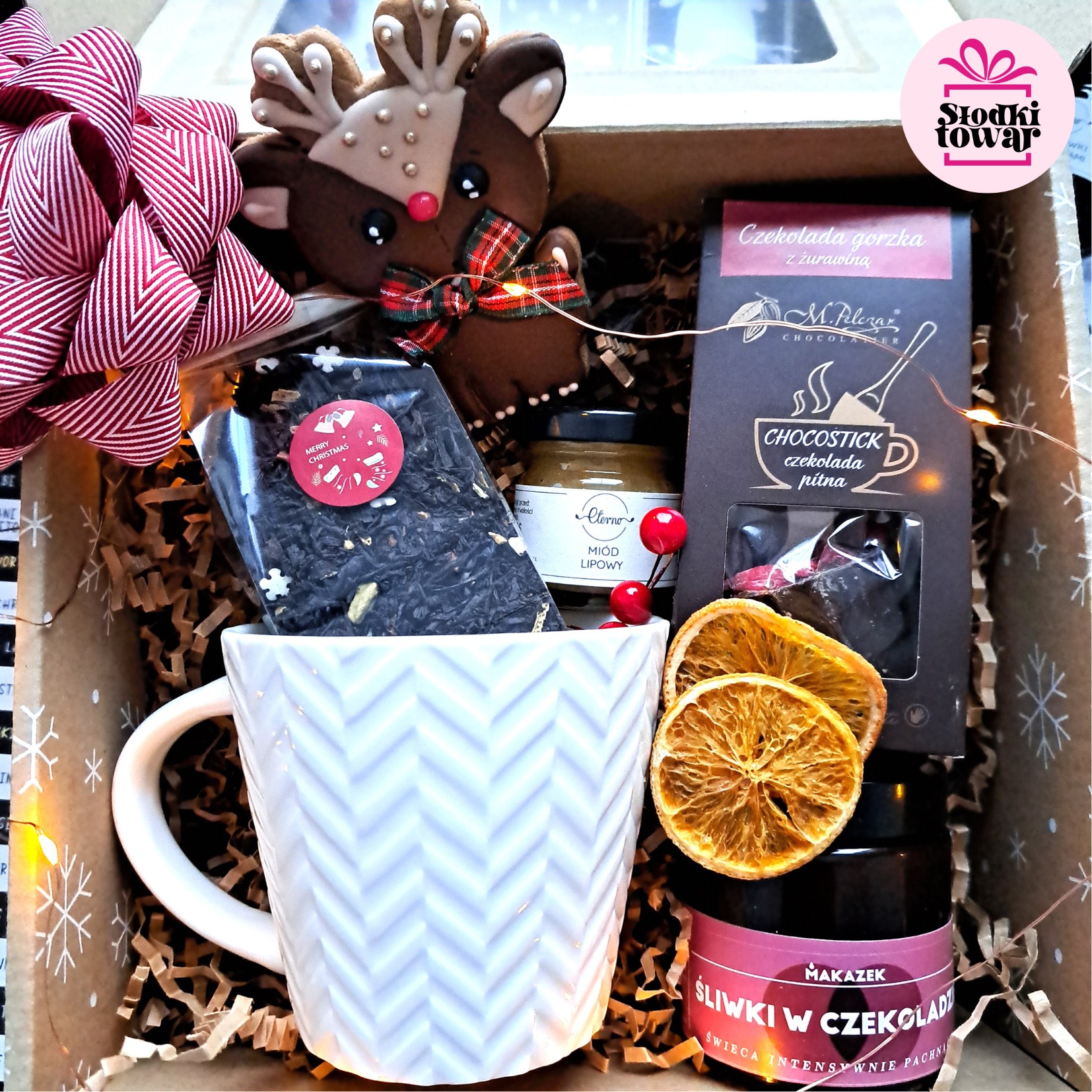 Świąteczny zestaw upominkowy w kartonowym pudełku z dekoracyjną kokardą, zawierający kubek z herbatą, miód lipowy, czekoladę pitną, suszone pomarańcze, piernik w kształcie renifera i świecę...