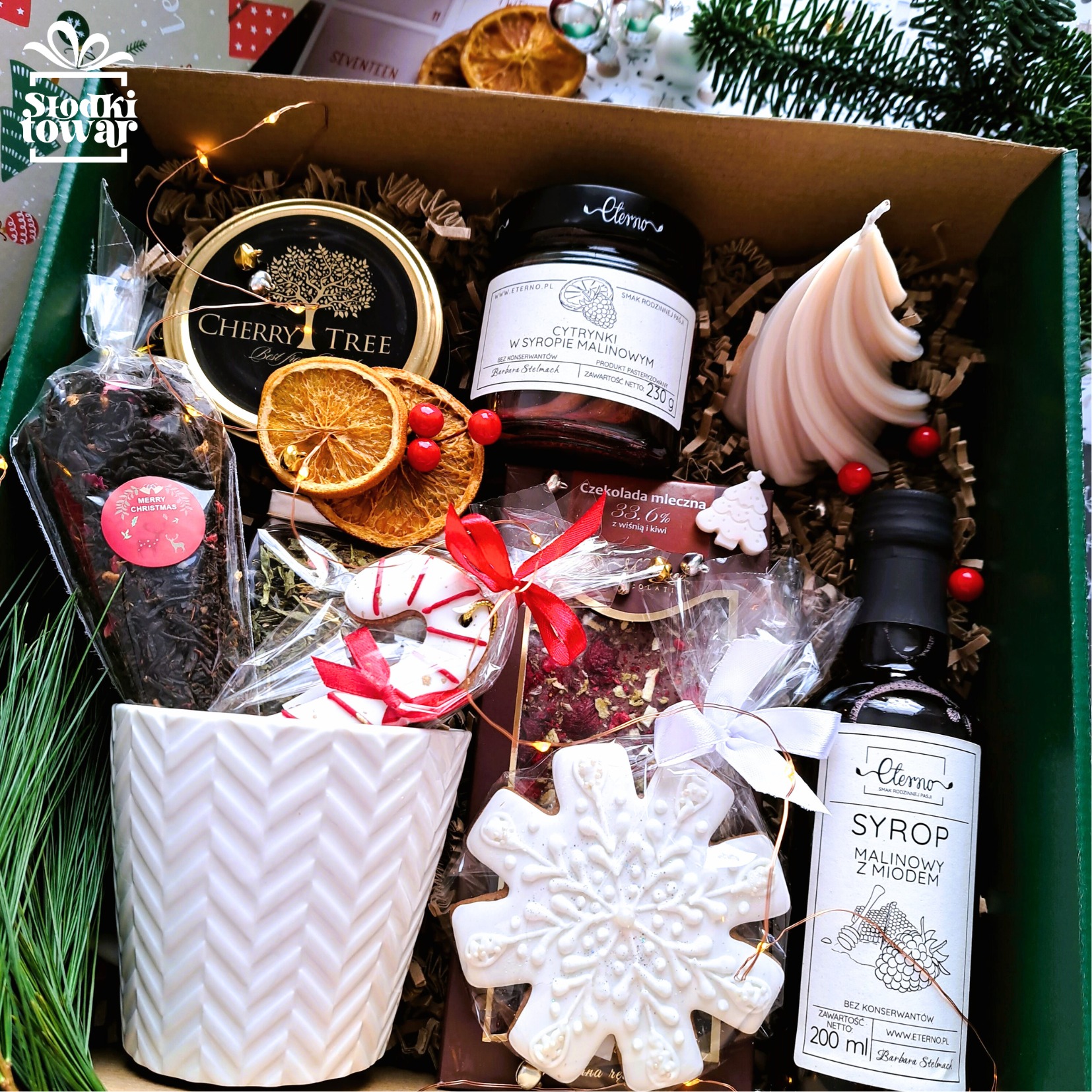 Świąteczny zestaw upominkowy w zielonym pudełku, zawierający herbatę w ozdobnym kubku, miód cytrynowy, syrop malinowy, czekoladę, piernikowe ozdoby i świeczkę w kształcie choinki, udekorowany...