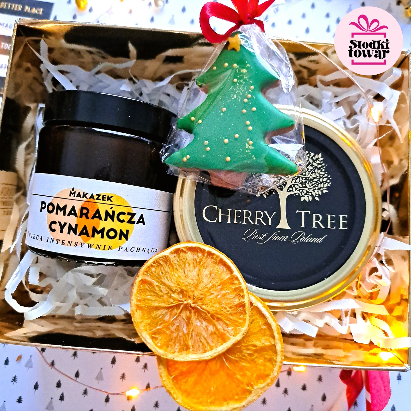 Zestaw upominkowy w złotym pudełku wypełnionym ozdobnym papierem, zawierający słoik z etykietą 'Pomarańcza Cynamon', ciasteczko w kształcie choinki z czerwoną kokardką, słoik z napisem 'Cherry...