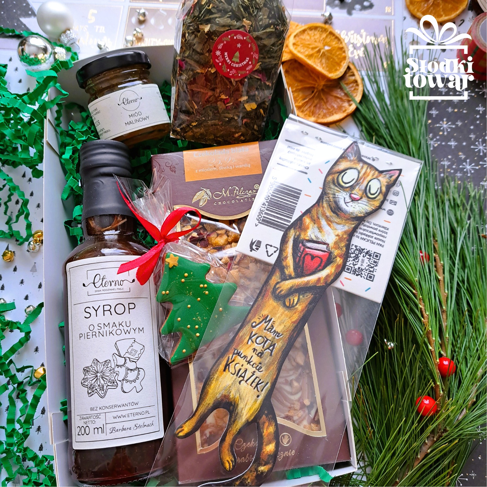 Zawartość kartonowego pudełka: miód, herbata, syrop piernikowy, czekolada, piernik w kształcie choinki i zakładka do książki z rysunkiem kota, całość udekorowana zielonymi serpentynami i gałązkami...
