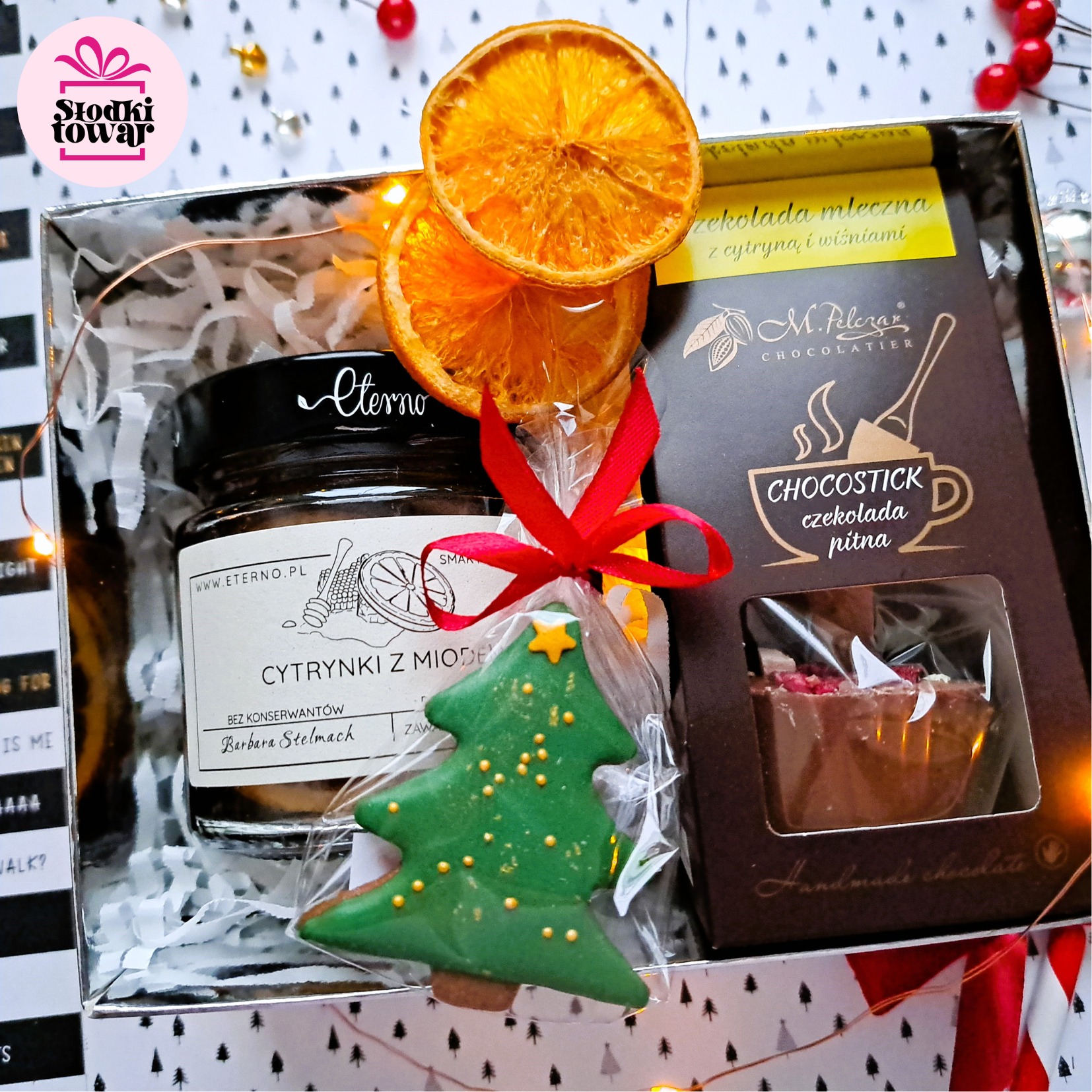 Świąteczny kosz prezentowy z miodem cytrynowym marki Eterno, czekoladą pitną M. Pelczar i piernikowym drzewkiem, ozdobiony suszonymi pomarańczami i czerwoną wstążką.