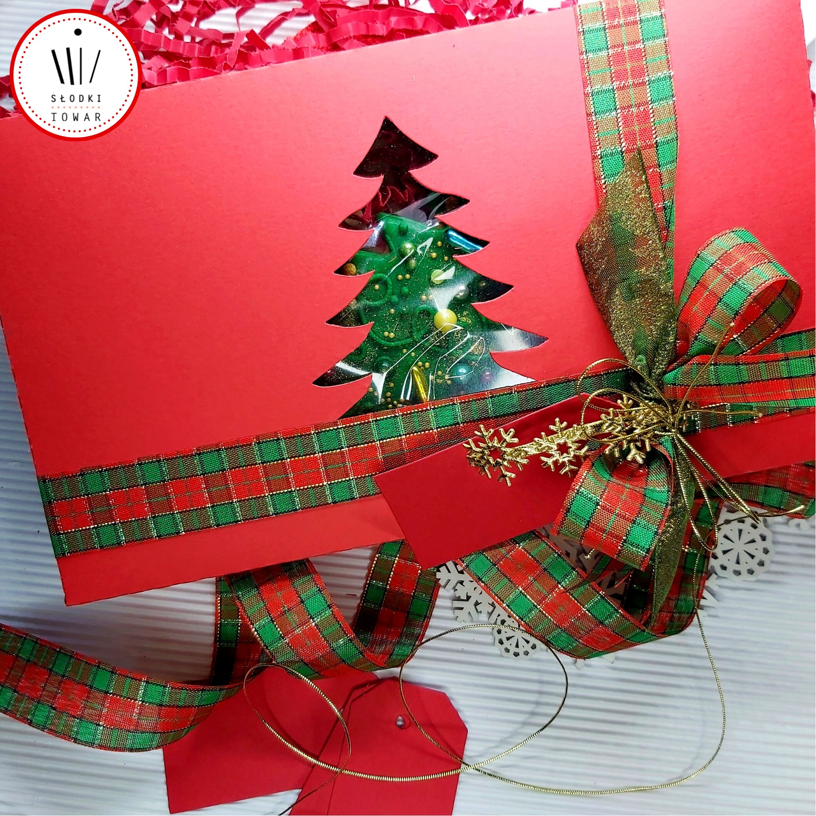 Czerwony kosz prezentowy z wyciętym kształtem choinki, wypełniony słodyczami, ozdobiony wstążką w kratę i złotymi śnieżynkami, oferowany w Gdyni.