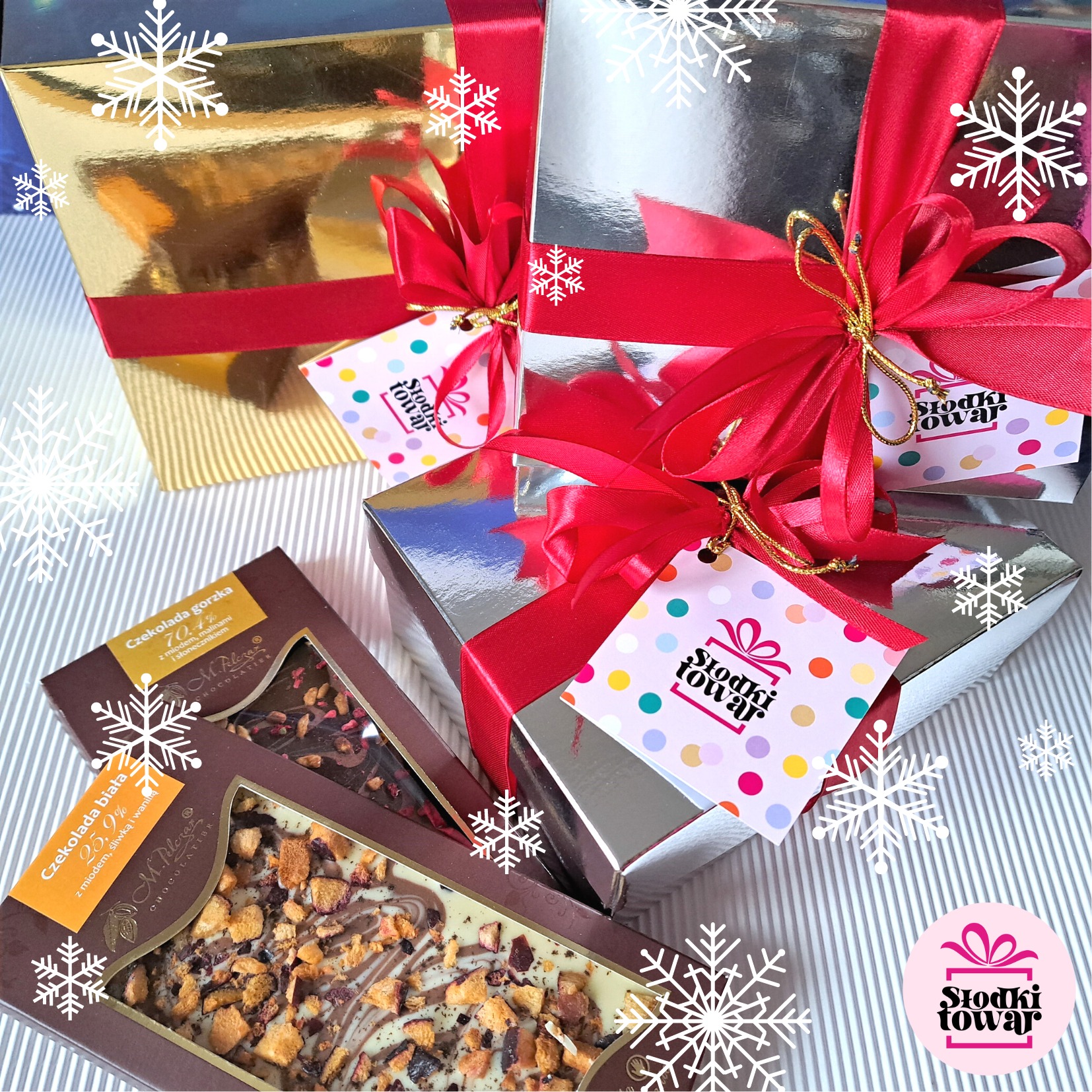 Dwa srebrne i jeden złoty błyszczący prezent zapakowany w pudełka przewiązane czerwoną wstążką, obok dwie otwarte czekolady rzemieślnicze z widocznymi dodatkami, całość na tle w biało-szare paski...