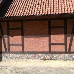 Michał Olechnowicz - Fragment ściany budynku z czerwonej cegły z elementami konstrukcji szkieletowej z ciemnego drewna, widoczny dach z dachówki ceramicznej i rynna, w tle drzewa.