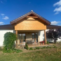 Michał Olechnowicz - Dom jednorodzinny z ciemnobrązowym dachem i drewnianą elewacją werandy, widok z ogrodu w słoneczny dzień. W tle błękitne niebo z lekkimi chmurami. Na pierwszym planie trawnik i elementy roślinności.